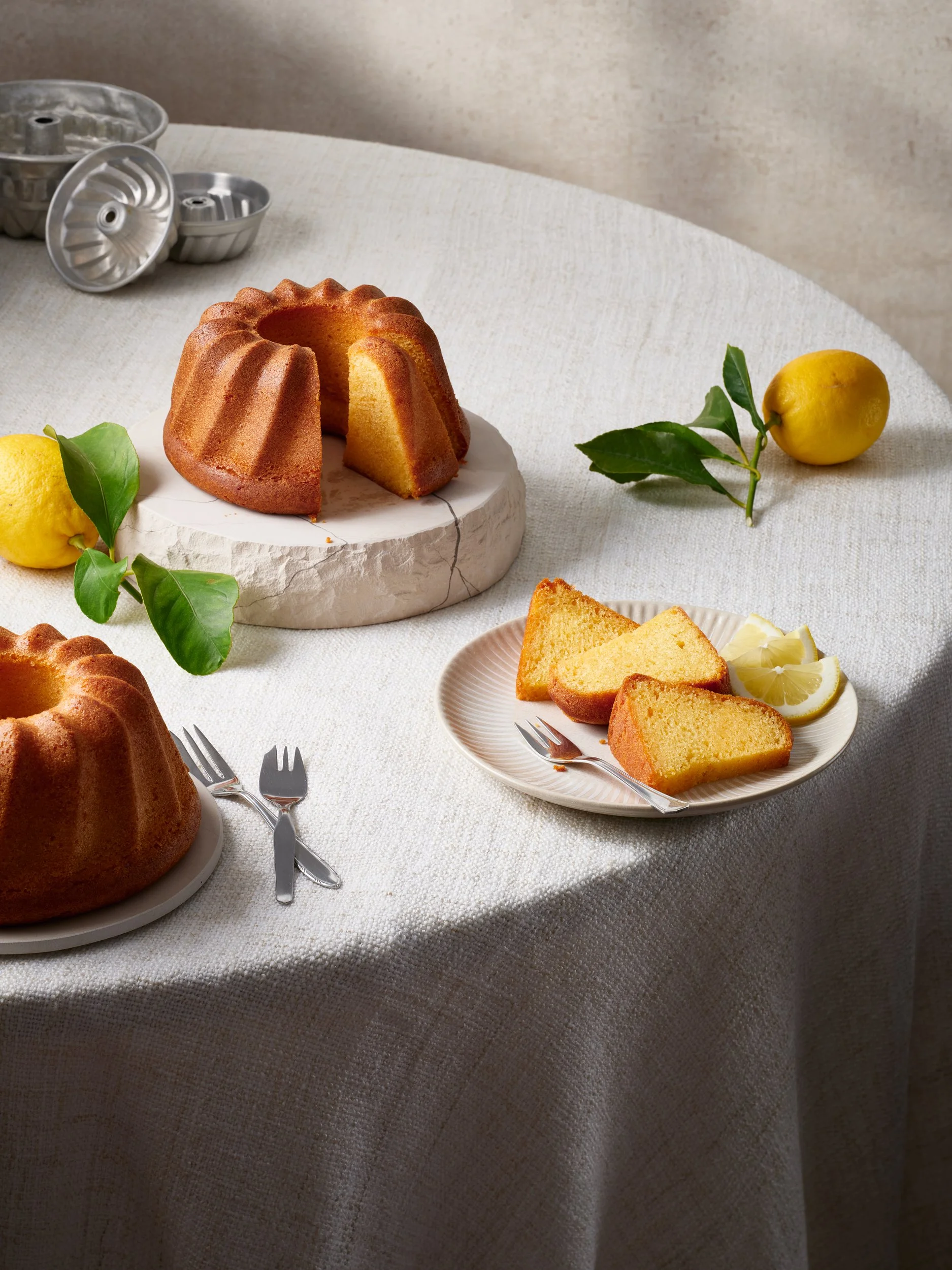 Ein gedeckter Tisch mit einem Gugelhupf, Zitronenscheiben, zwei Zitronen, grünem Blatt, einem Kastenformkuchen und Backformen im Hintergrund.