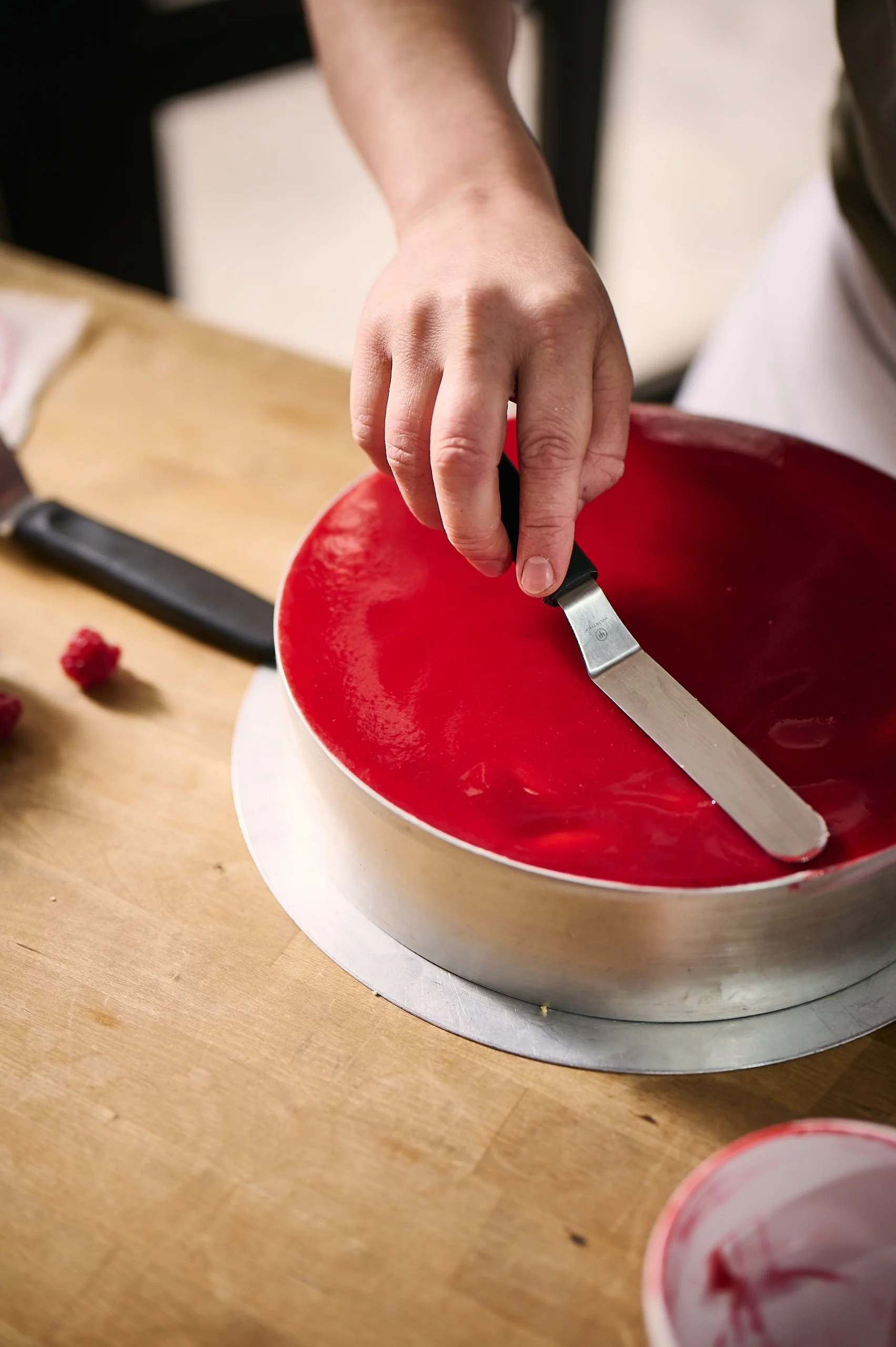 Eine Person glättet mit einem Spatel eine rote, glänzende Quarktorte auf einer runden Tortenplatte, während im Hintergrund eine Holztischplatte sichtbar ist.