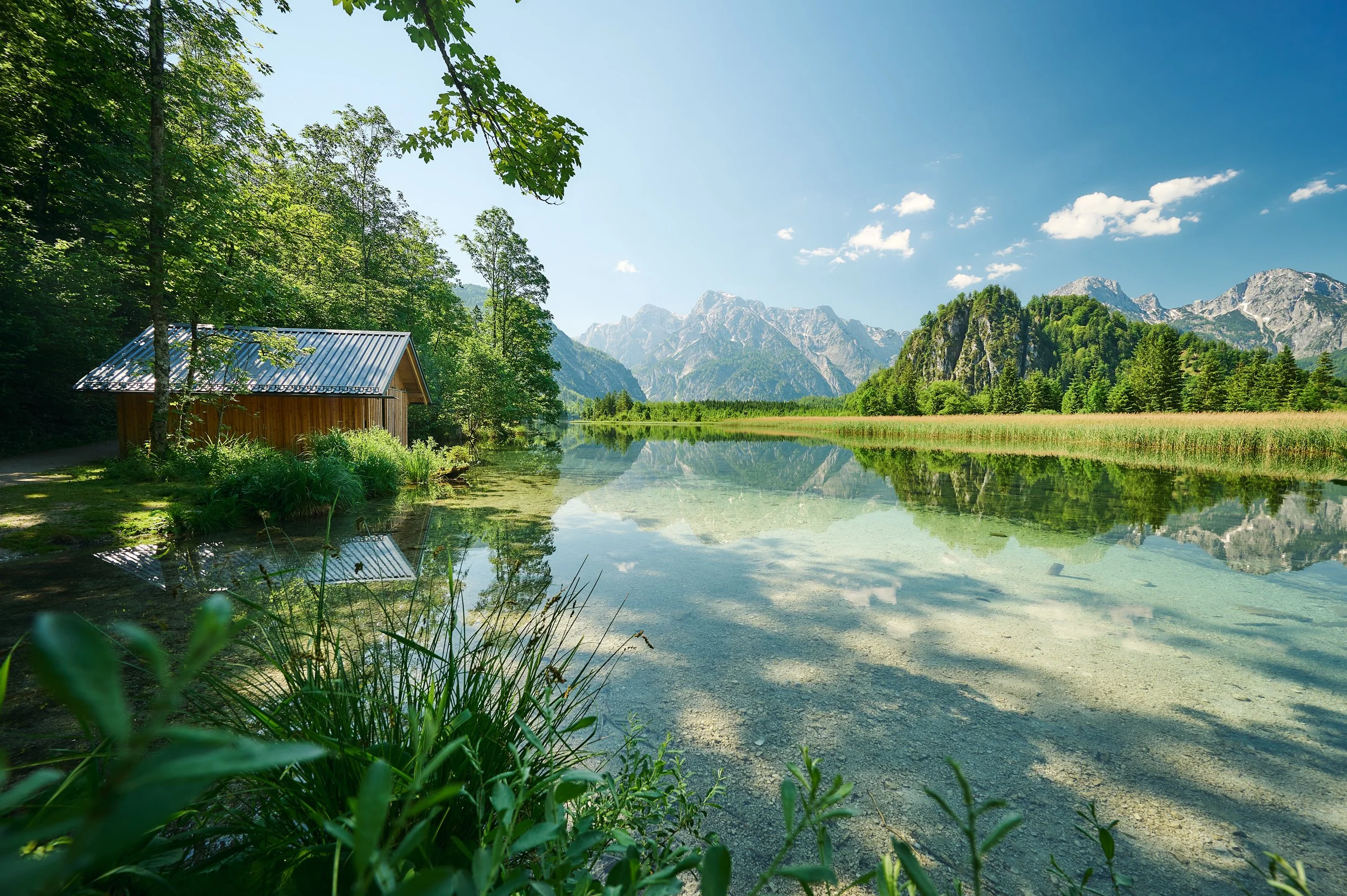 Ein ruhiger See umgeben von grünen Bäumen und hohen Bergen im Hintergrund, mit einem kleinen Holzhaus am Ufer, sonniges Wetter und blauer Himmel.
