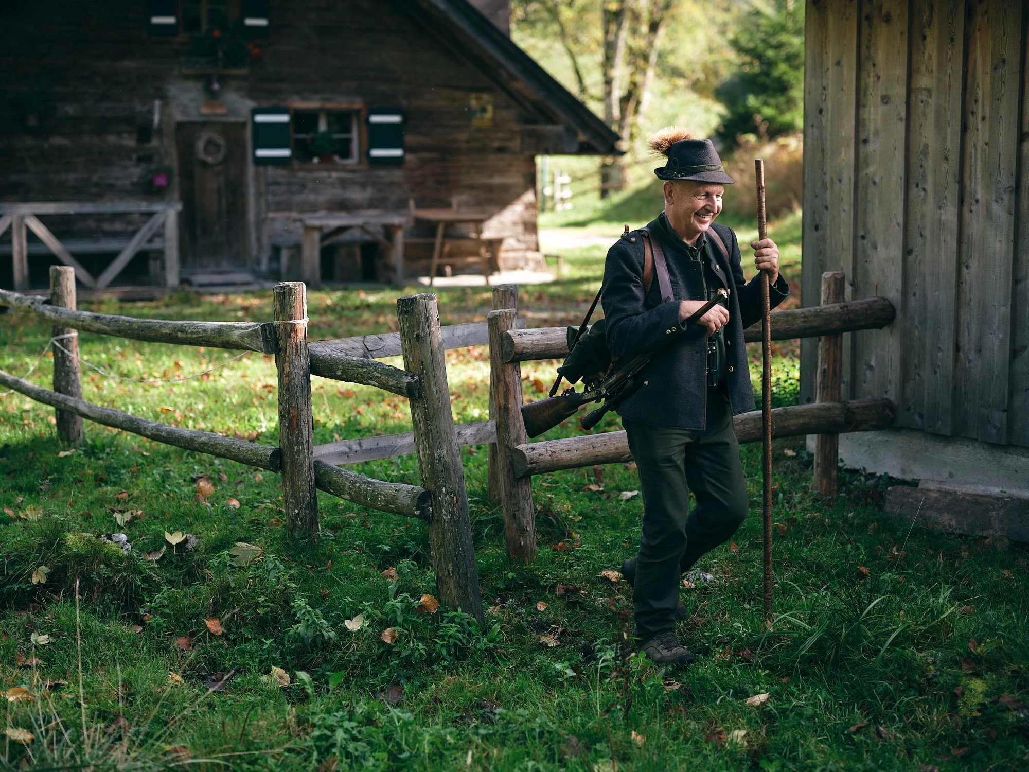 Ein älterer Mann in traditioneller Tracht mit Filzhut, der lachend einen Spazierstock hält, steht vor einer Holzzaun an einem ländlichen Holzhaus in einer grünen, herbstlichen Umgebung.