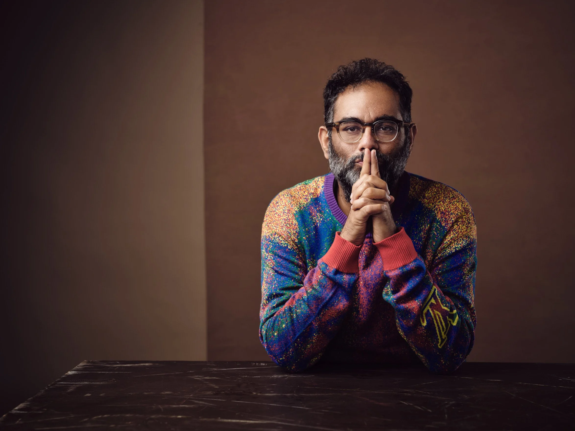Mann mit Bart und Brille, trägt einen bunten Pullover, sitzt nachdenklich an einem Tisch vor dunklem Hintergrund.