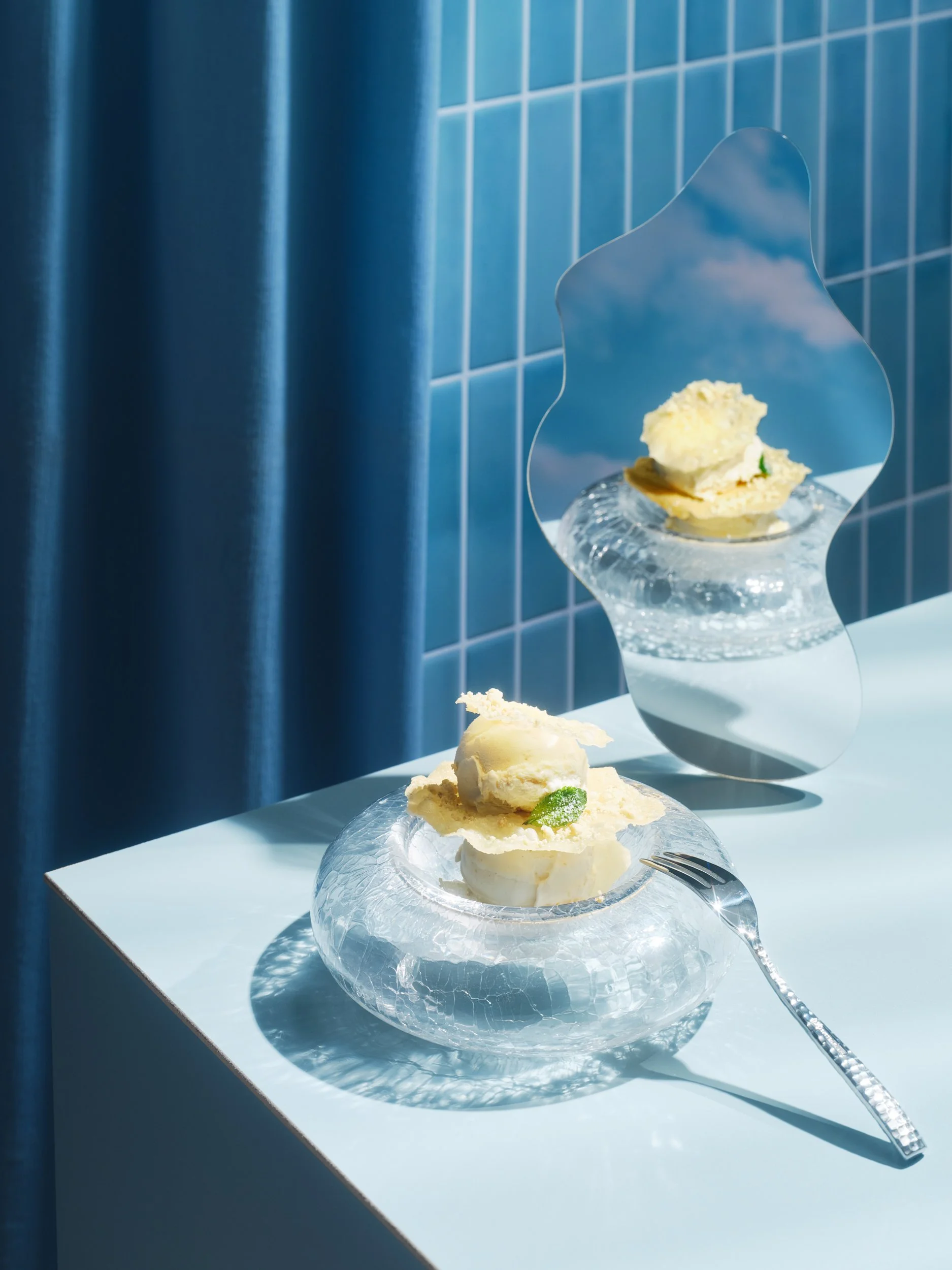 Glas Teller mit Vanilleeis und Apfelstrudelscheiben, serviert auf einem weißen Tisch vor blauer Fliesenwand, mit einer Gabel.