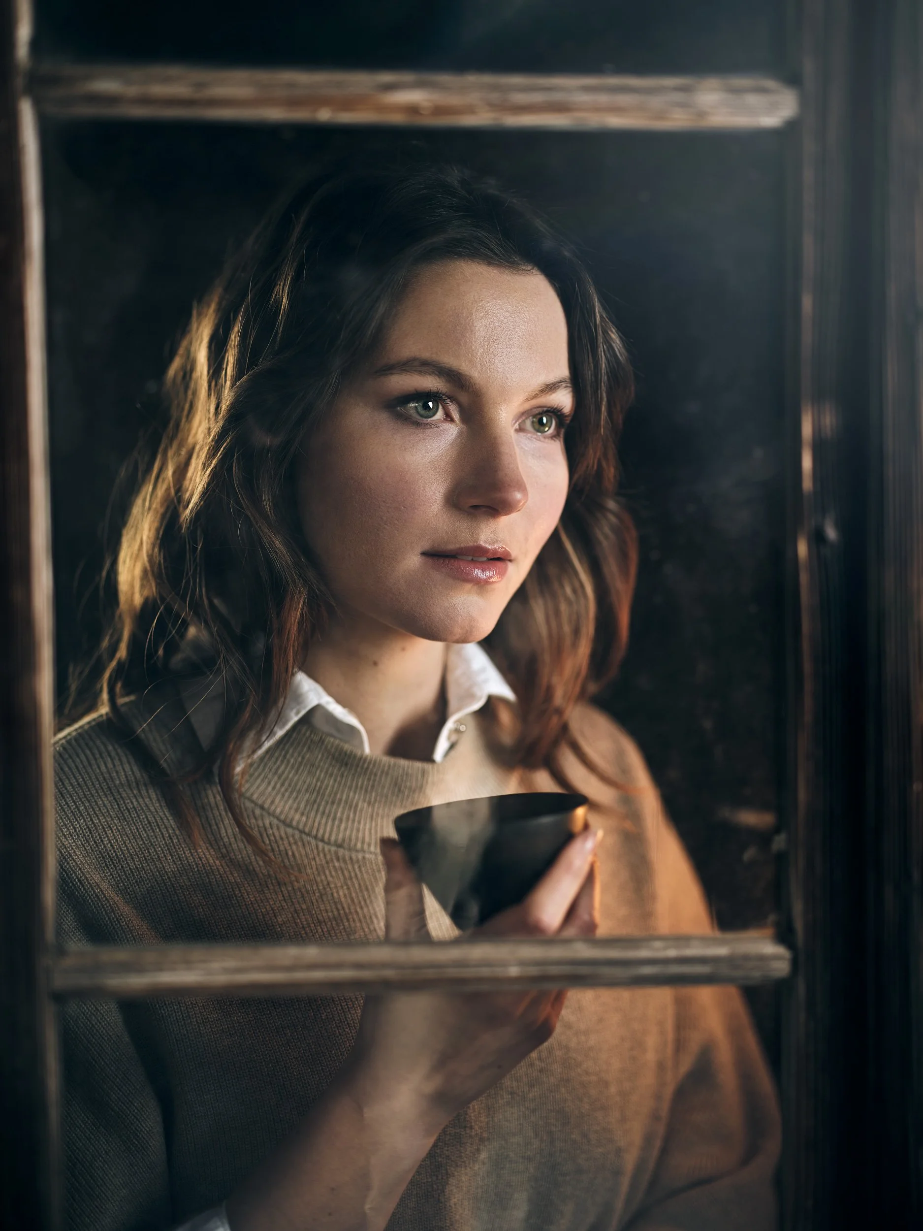 Frau mit braunen Haaren, die aus einem Fenster schaut und eine Tasse hält.