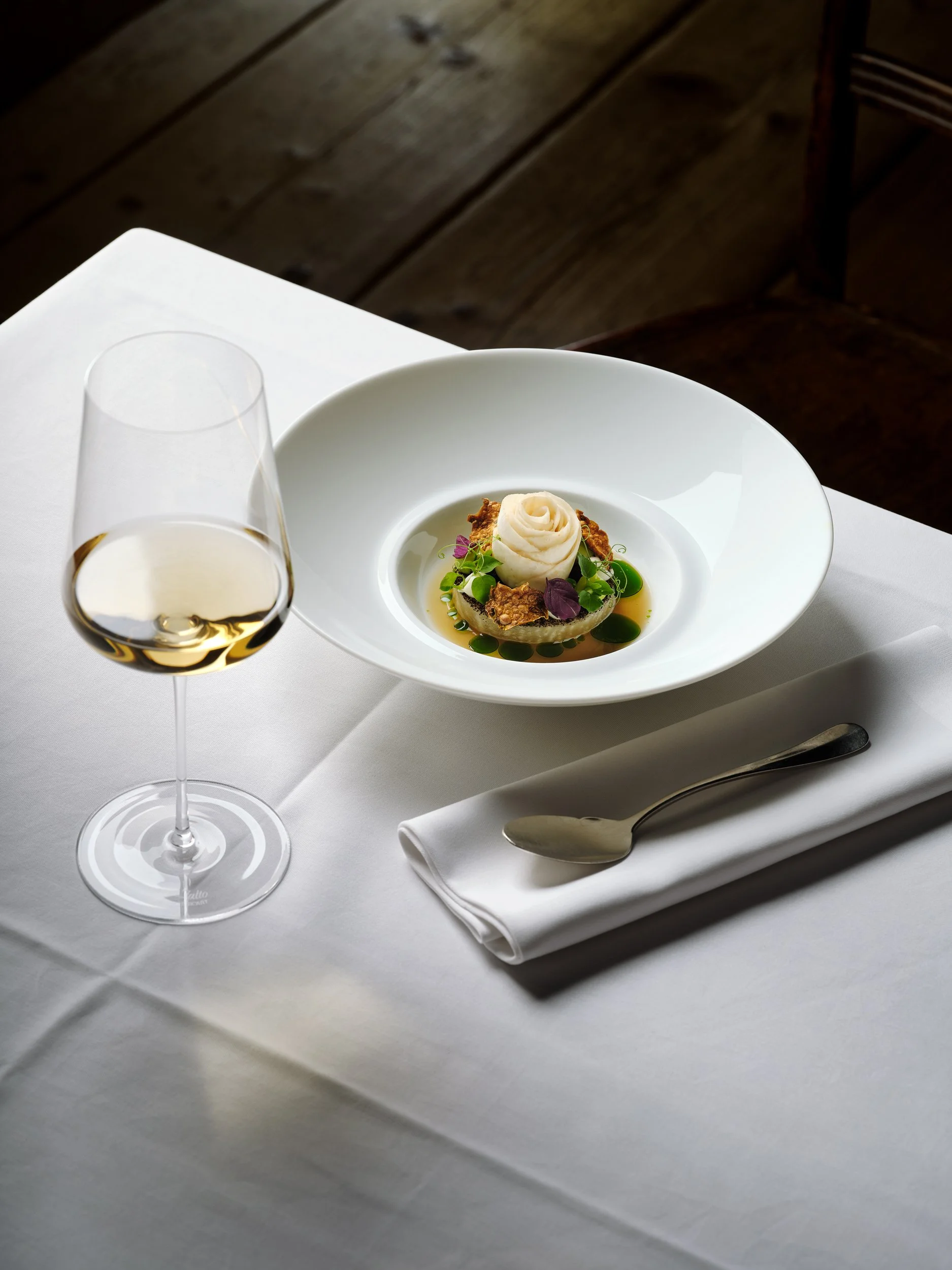 Gourmetgericht auf weißem Tisch: Ein kunstvoll angerichteter Teller mit einer kleinen Speise, umgeben von grünen und lila Garnierungen. Ein Glas Weißwein und ein Löffel auf einer Serviette daneben.