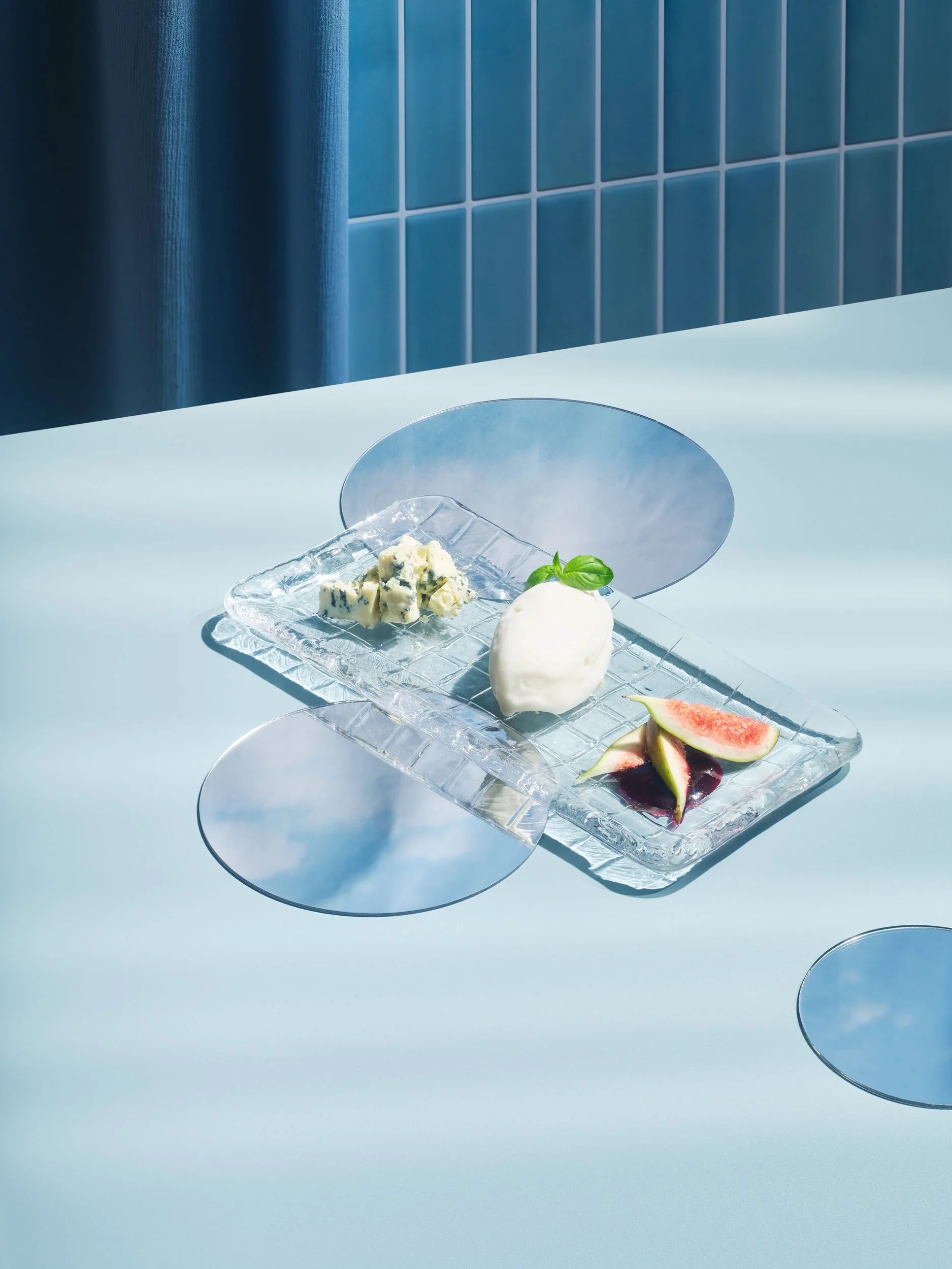 Ein Teller mit Blauschimmelkäse, Feigen, einer Kugel Eis, Feigensauce und einer Basilikumblatt auf einem Glas-Servierteller auf einem weißen Tisch, im Hintergrund blaue Fliesen.