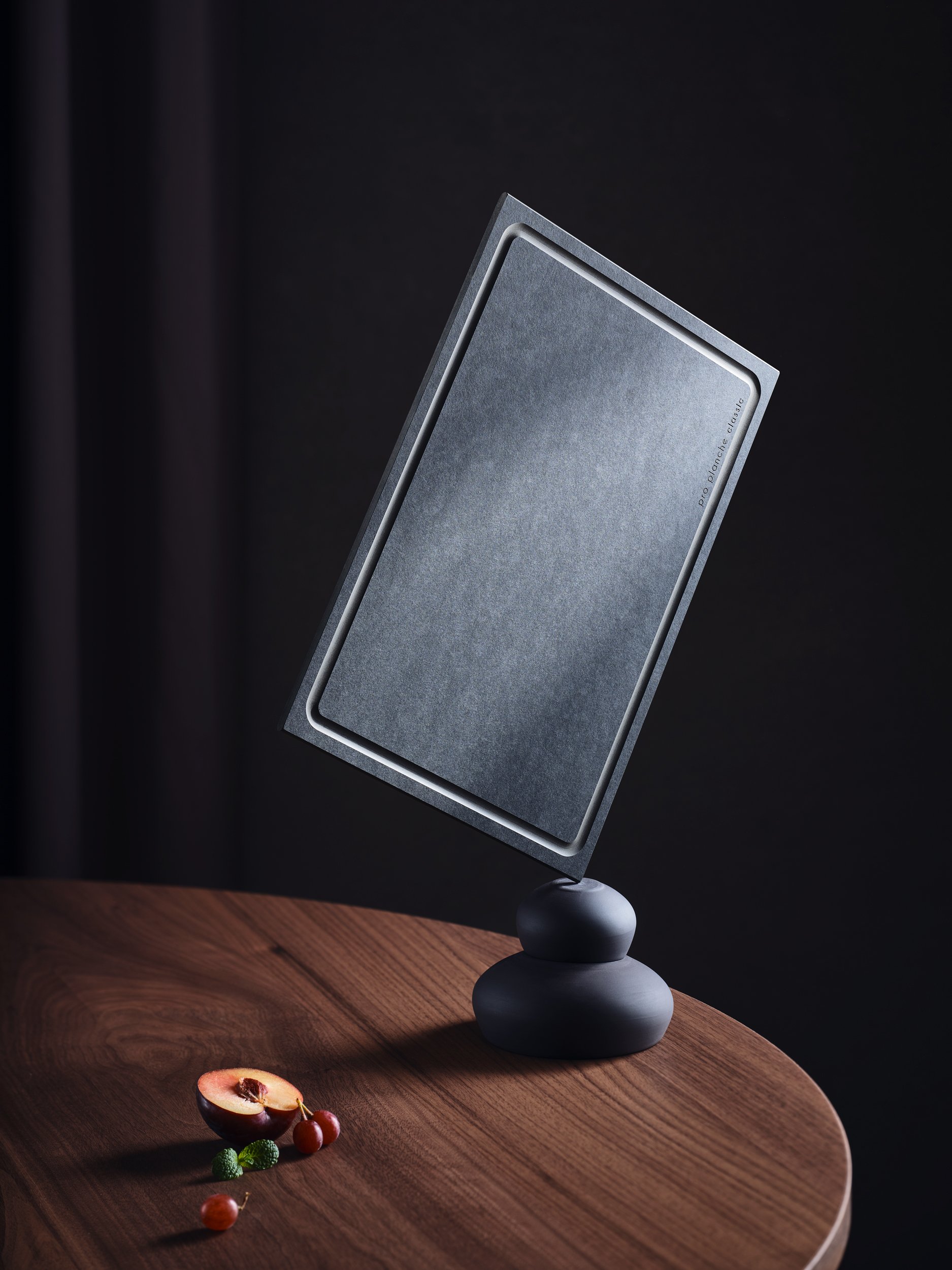 Ein modernes, dunkles Tablett auf einem Holz Tisch mit einem Prop, das wie ein schwebendes Tablet aussieht, und ein paar Früchte, darunter eine aufgeschnittene Pflaume, auf dem Tisch.