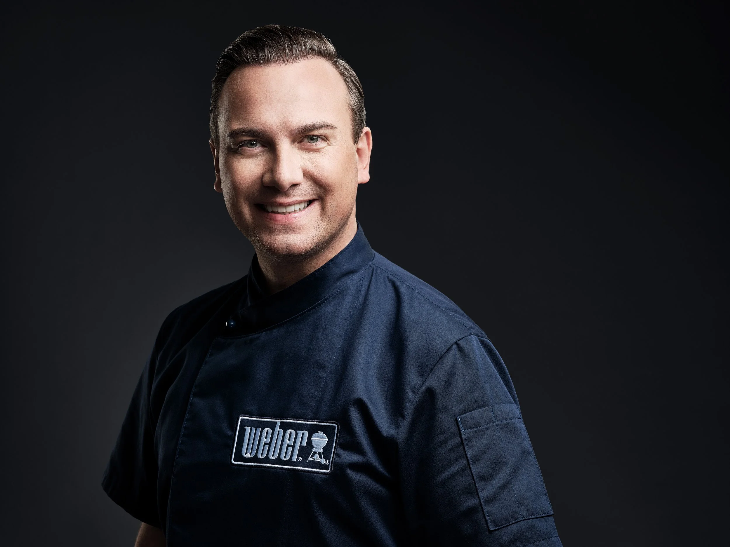 Ein Mann in einer dunklen Uniform mit einem Weber-Logo, vor einem schwarzen Hintergrund, lächelt in die Kamera.