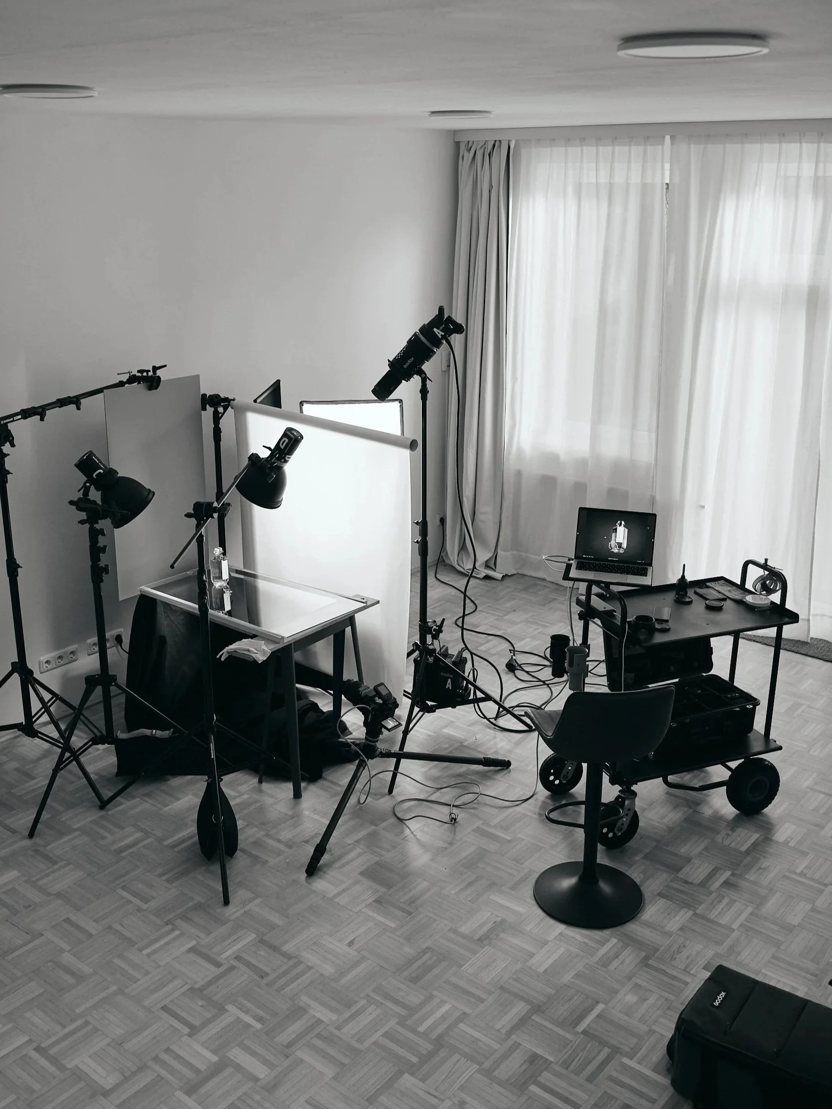 Ein Fotostudio mit verschiedenen Kameraausstattungen, Beleuchtung, Monitor und einem Laptop vor einer weißen Wand mit Vorhängen.