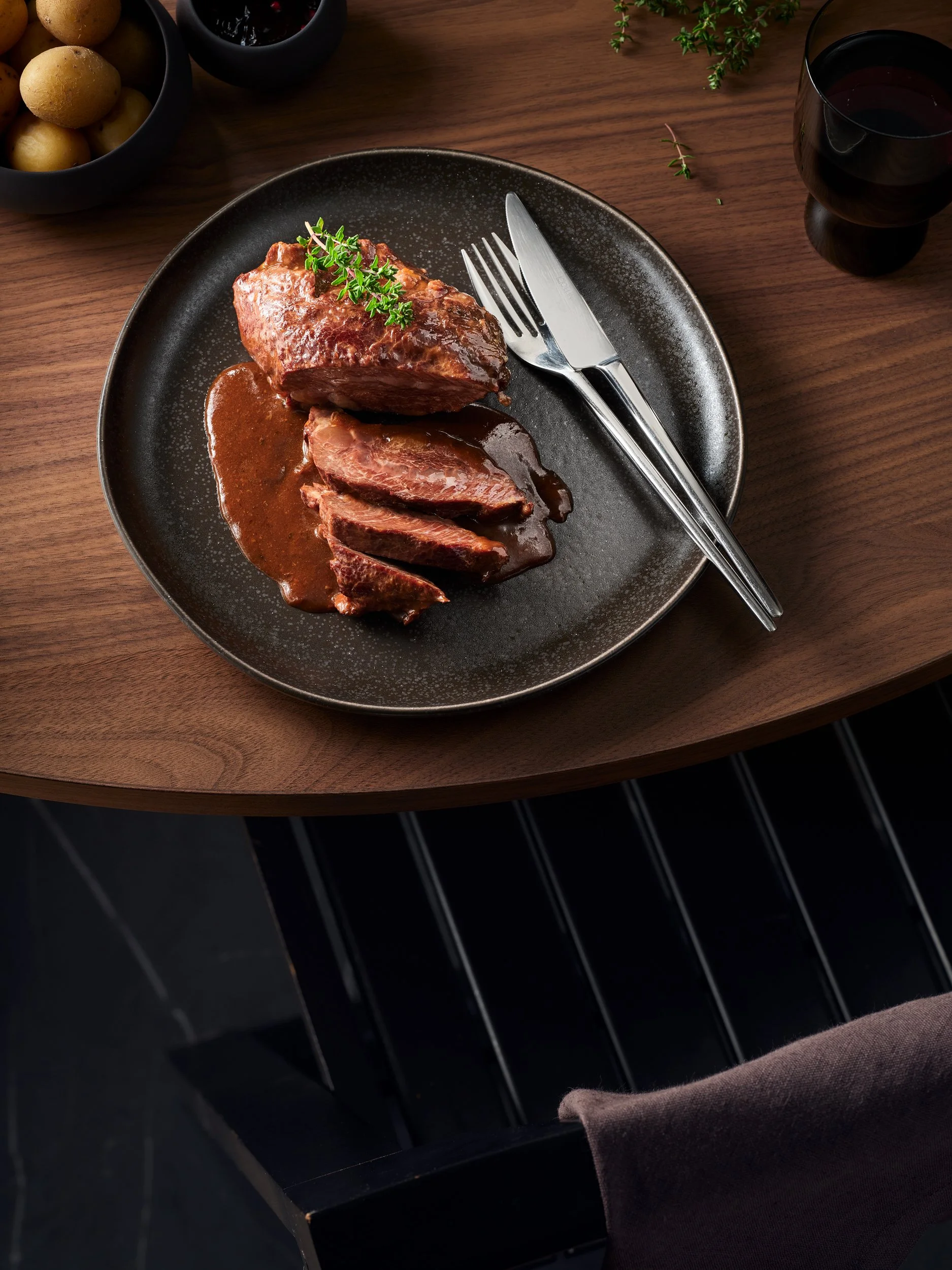 Ein Teller mit rosa gebratenem Steak, in Scheiben geschnitten, auf einem schwarzen Teller, mit einer kleinen grünen Kräuter Garnitur, auf einem Holztisch, daneben ein Messer und eine Gabel, im Hintergrund ein Glas Rotwein, und zwei schwarze Schalen mit Kartoffeln und einer dunklen Sauce.
