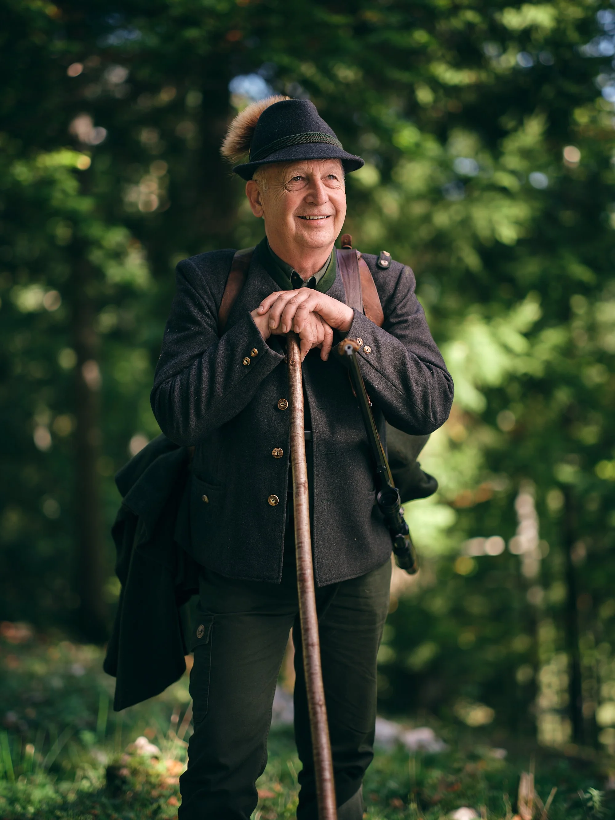 Ein älterer Mann steht im Wald, trägt einen traditionellen Hut mit Edelweiß und sitzt auf einem Spazierstock, lächelt. Er trägt eine grüne Jacke und eine dunkle Hose.