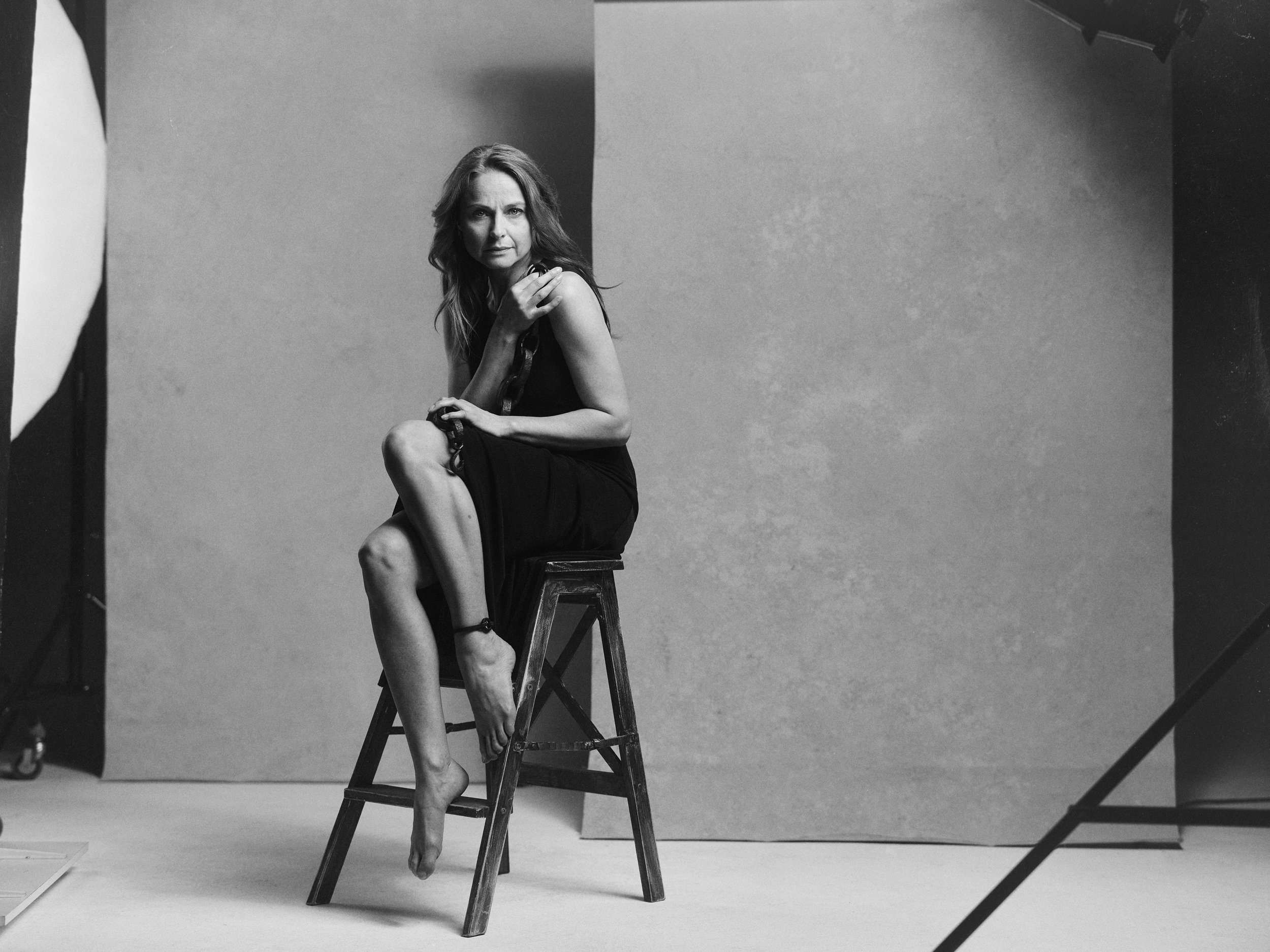 Eine Frau sitzt auf einer hölzernen Leiter in einem Fotostudio. Sie trägt ein schwarzes Kleid, hat langes Haar und schaut direkt in die Kamera. Der Hintergrund ist einfarbig grau.
