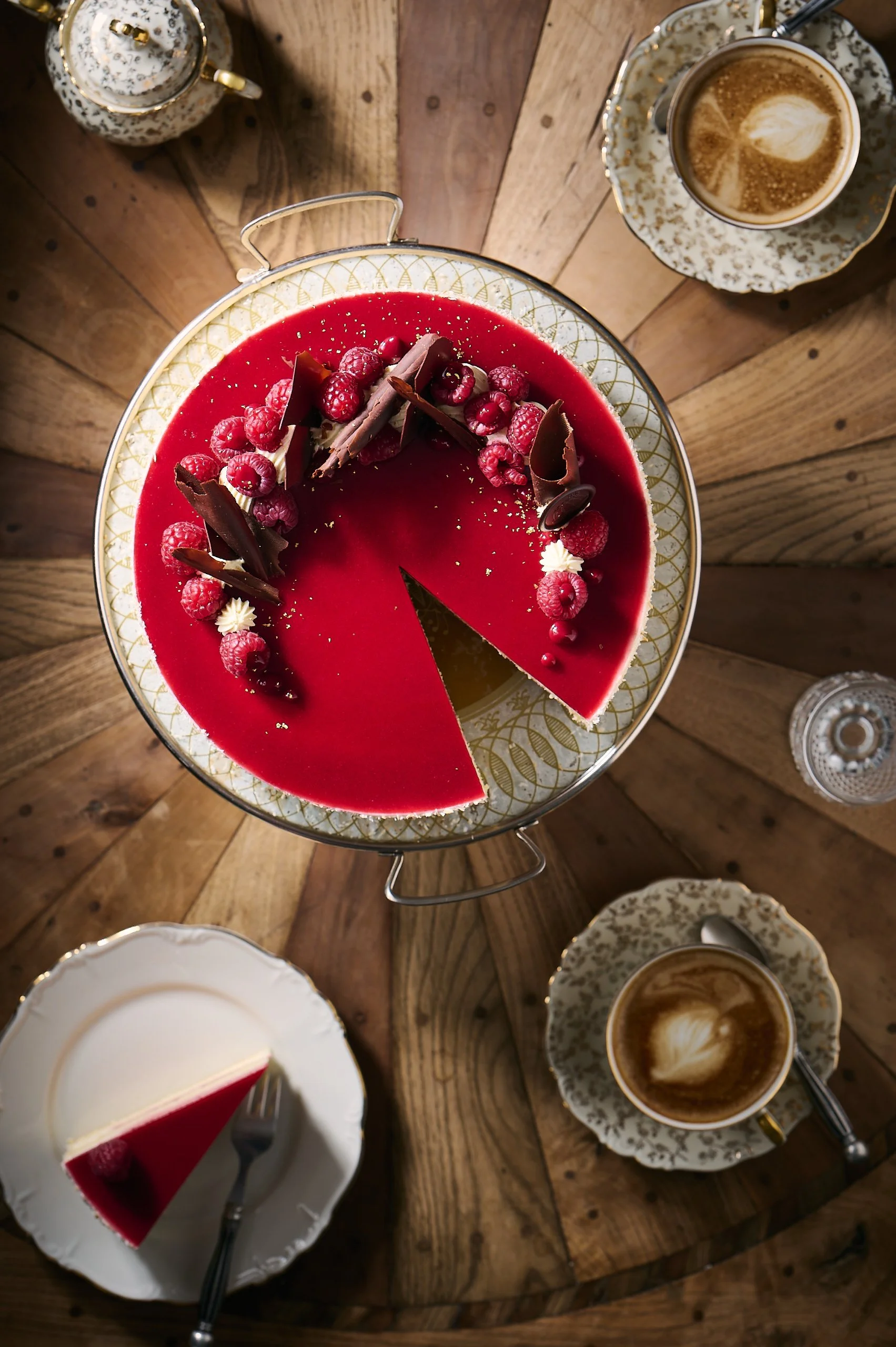 Ein runder Tisch mit einer roten Obsttorte mit Himbeeren, Schokoladenspänen und Goldstaub, begleitet von zwei Tassen Kaffee mit Milchschaum. Das Bild ist von oben aufgenommen.