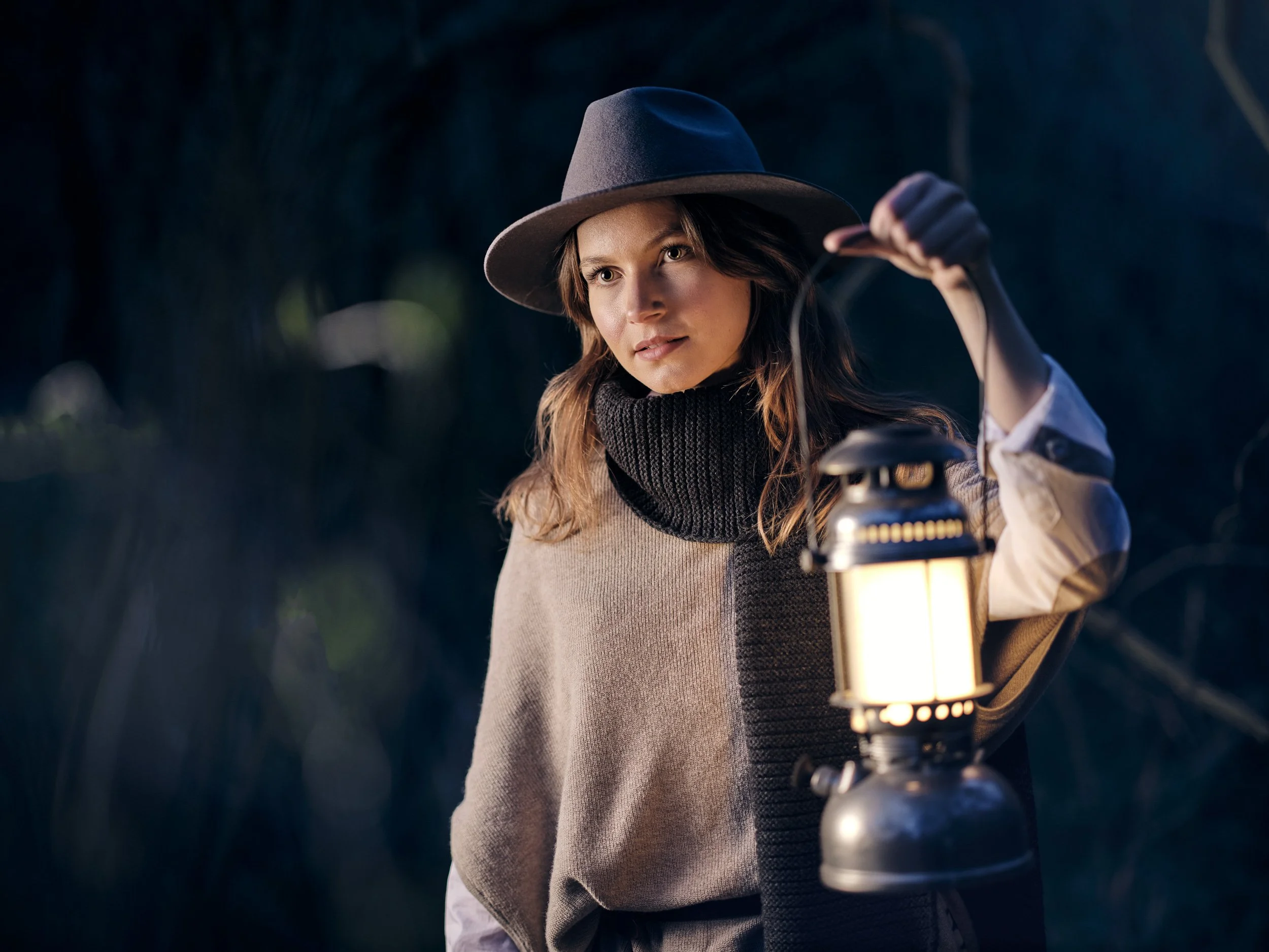 Frau im Wald mit Laterne und Hut, trägt Schal und Poncho.