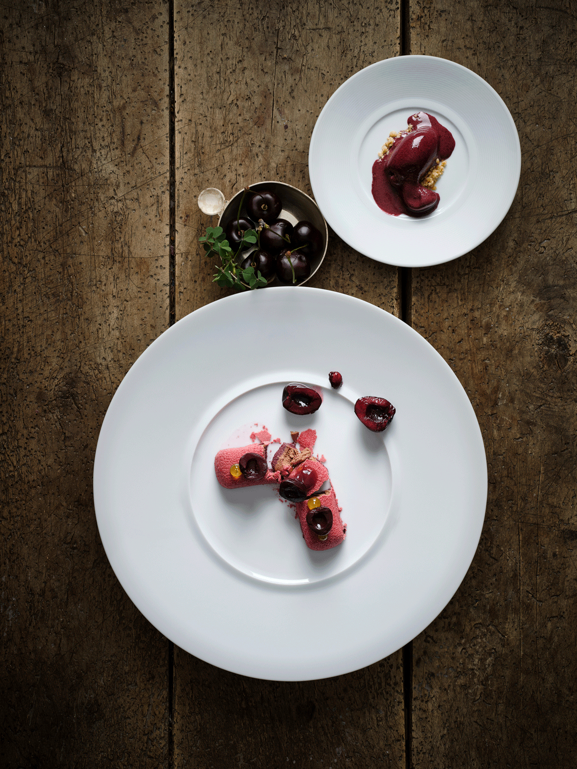 Dessert auf einem weißen Teller, mit Kirschen, Creme und Dekoration, daneben eine Schüssel mit frischen Kirschen, auf einem Holztisch.