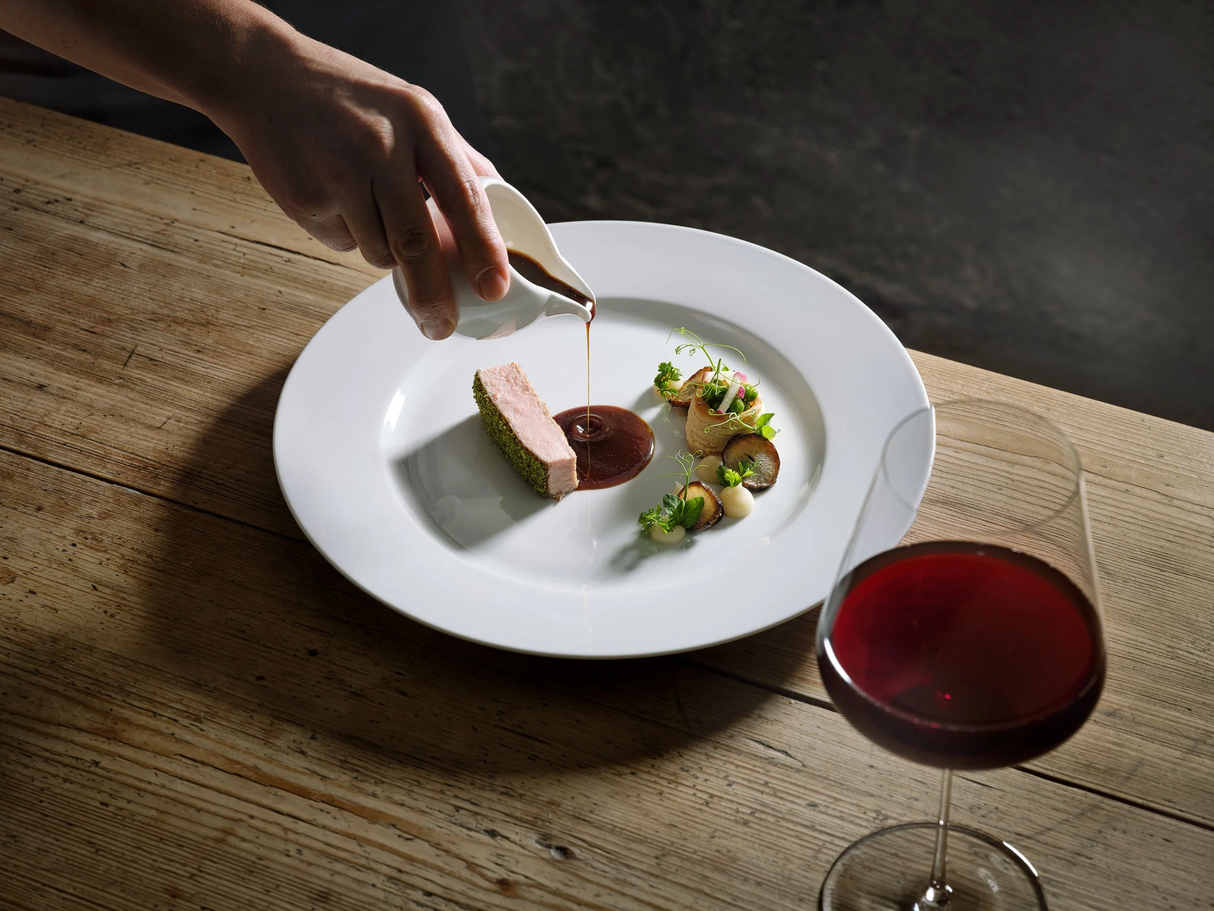 Ein Teller mit einem Stück Fleisch, das mit einer dunklen Soße übergossen wird, und einer kunstvoll angerichteten Beilage aus Gemüse. Daneben ist ein Glas Rotwein sichtbar. Eine Hand gießt die Soße auf den Teller.