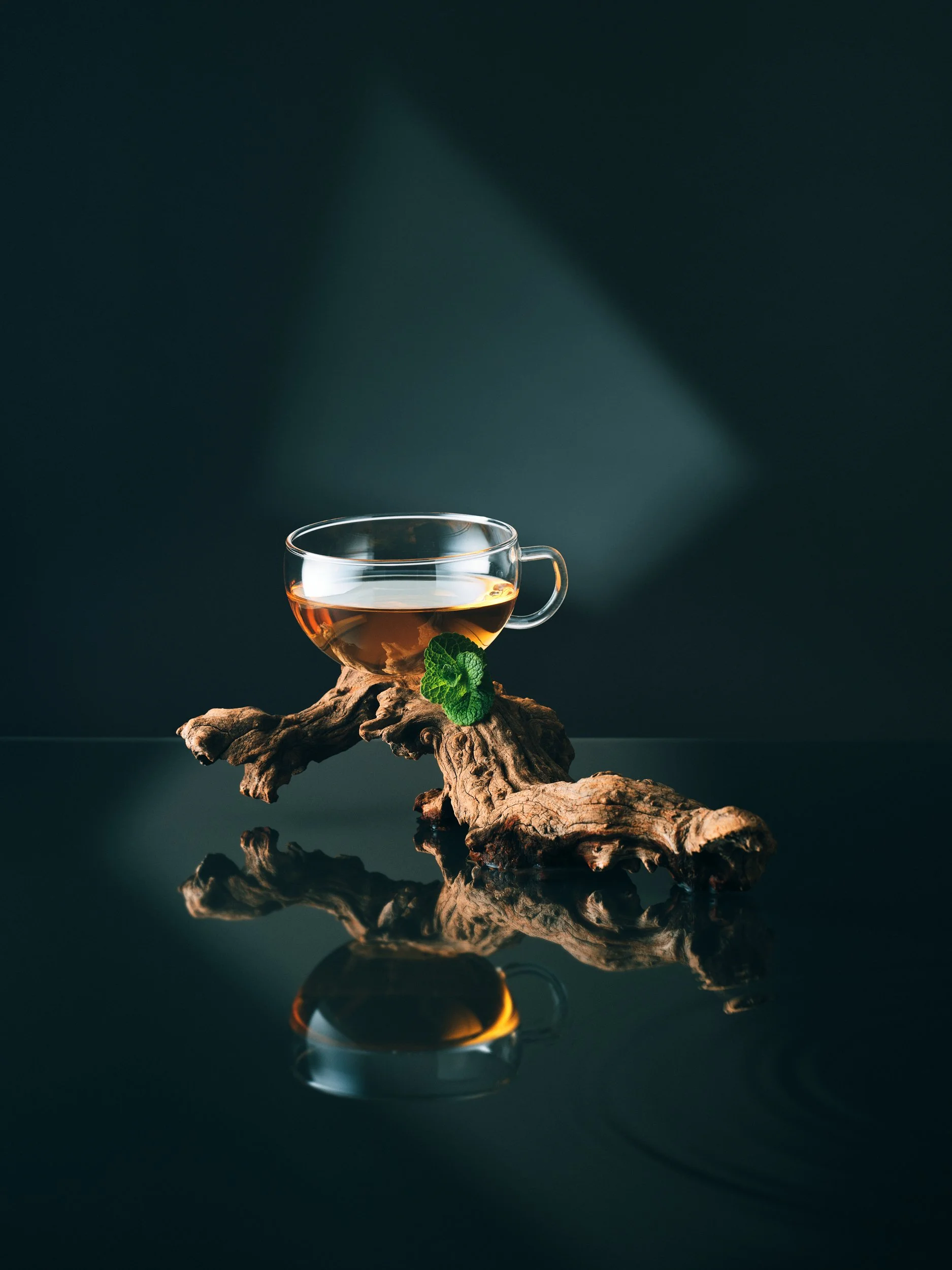 Eine Teetasse mit Kräuterteem auf einem stilvollen Holzkohle- oder Wacholderholzstück, mit einem grünen Minzblatt neben der Tasse vor einem dunklen Hintergrund.