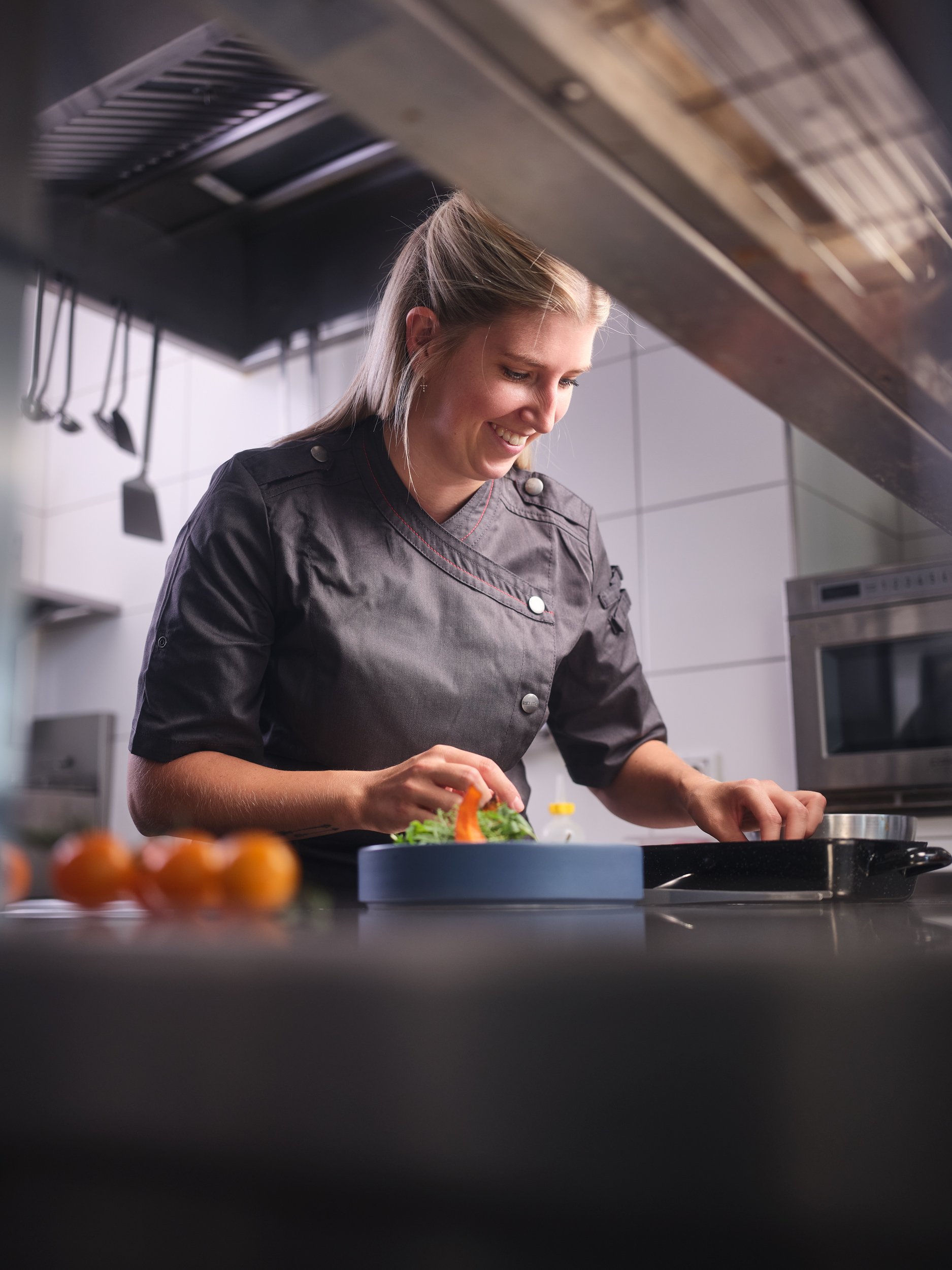 Eine junge Frau in einer schwarzen Kochjacke bereitet in einer modernen Küche Essen vor, während sie lächelnd auf die Kochplatte schaut.