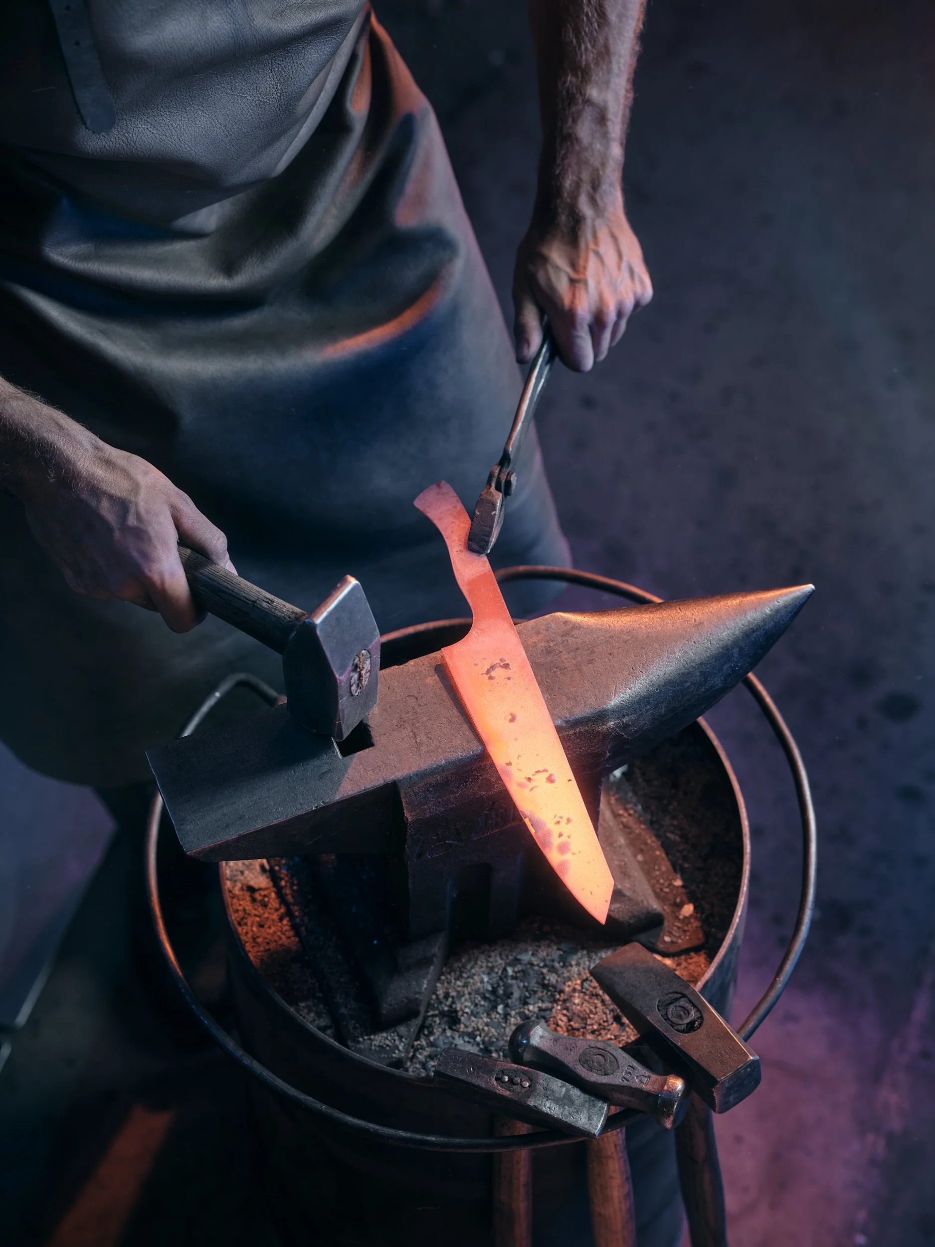 Schmied, der ein glühendes Metallstück auf einem Amboss bearbeitet, mit einem Hammer und einer Zange in der Hand, umgeben von Schmiedewerkzeugen.
