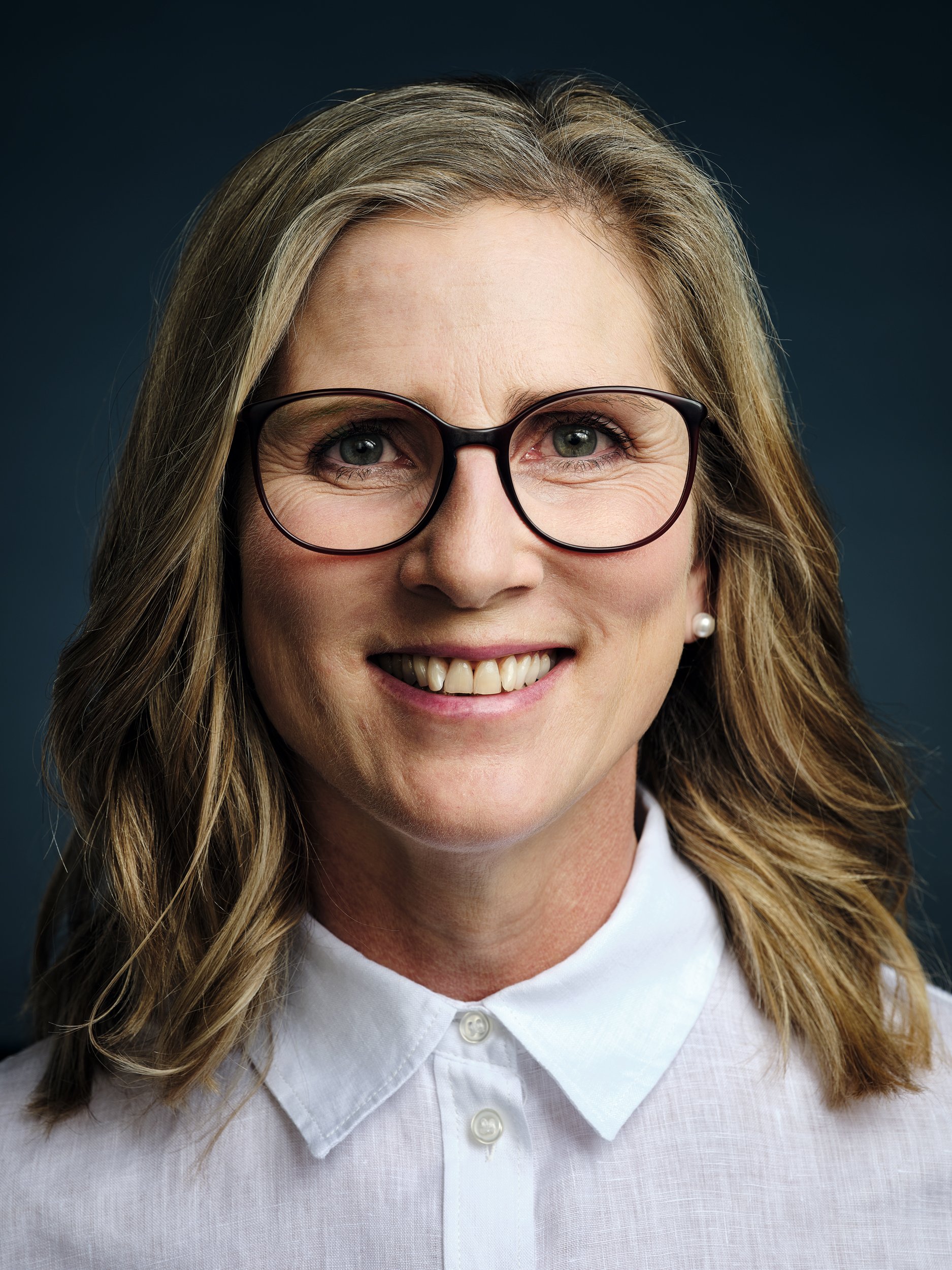 Nahaufnahme eines lächelnden Frau mit Brille, braunem Haar und weissem Hemd vor dunklem Hintergrund.