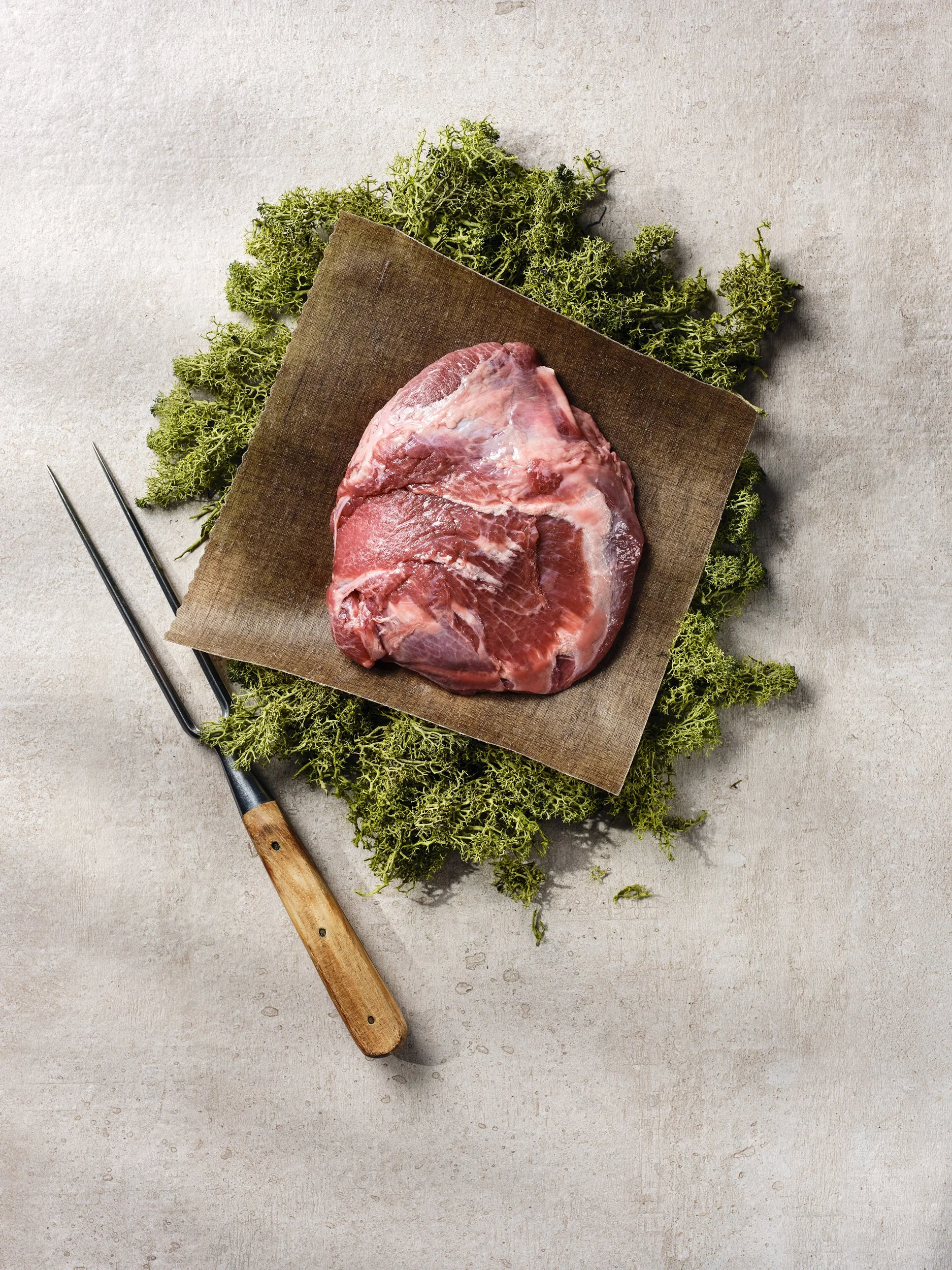 Rohes Fleisch, auf einer Steinplatte mit frischen Kräutern und einer Fleischgabel daneben arrangiert.