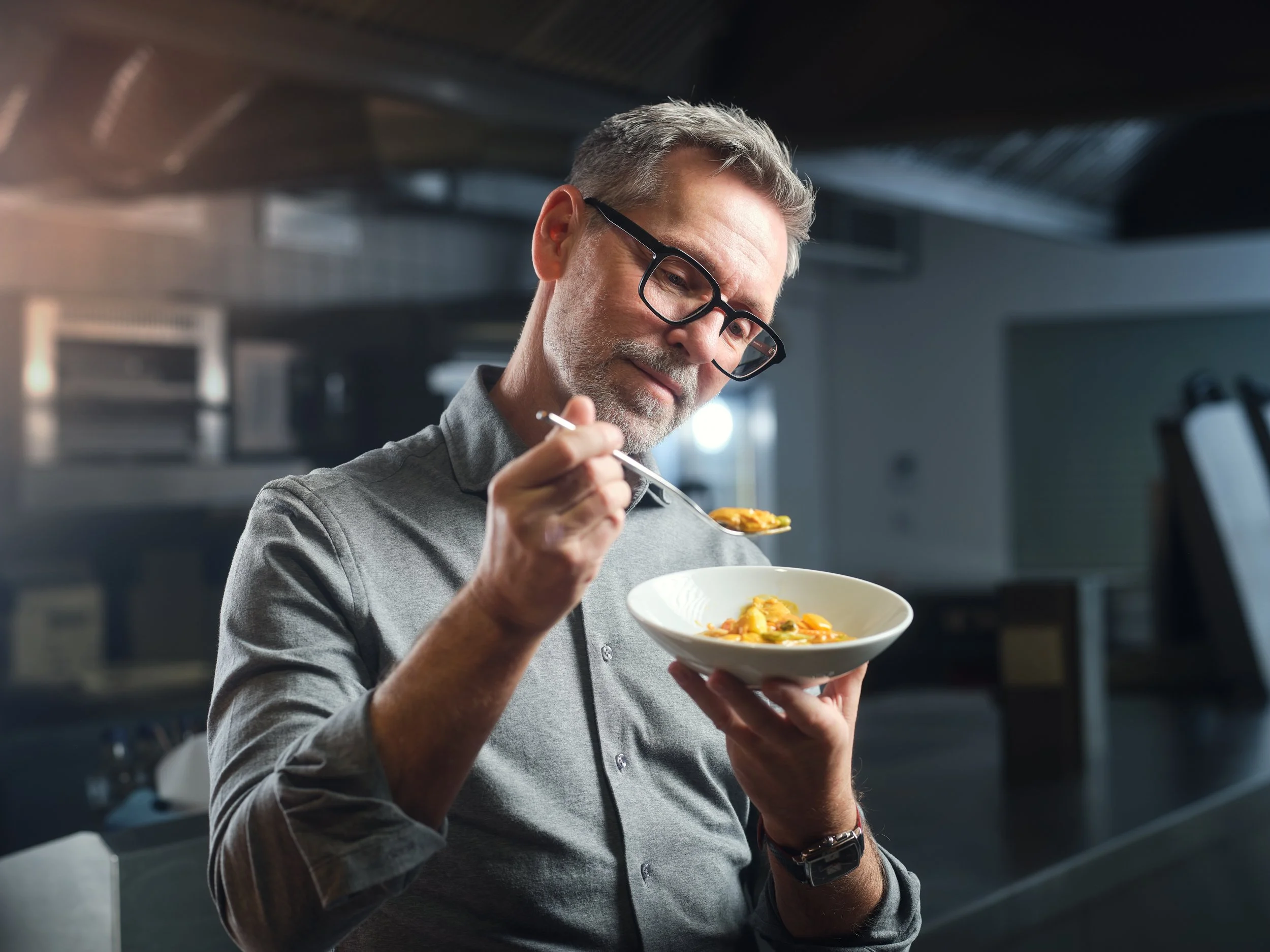 Ein älterer Mann mit Brille und grauem Haar isst eine Schüssel mit Pasta in einer modernen Küche.