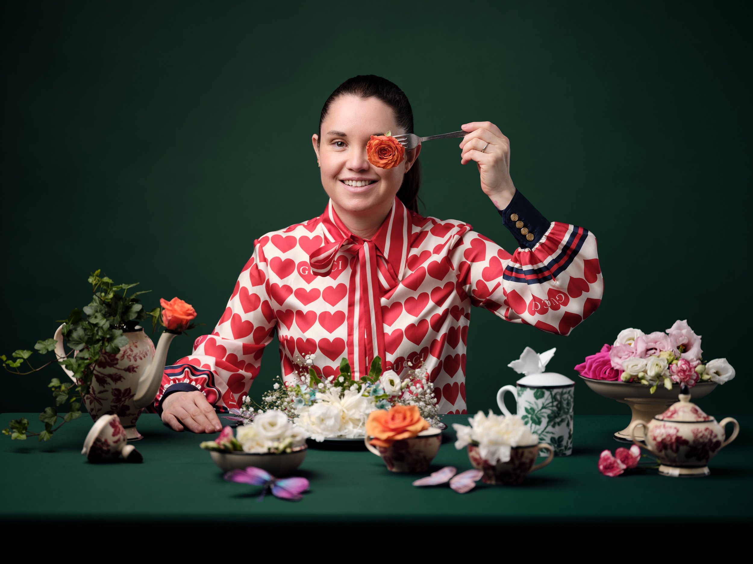 Frau in Herzmuster-Bluse hält eine orange Blume mit einer Gabel vor Auge, sitzt an Tisch mit Blumenschmuck, Teekanne und Tassen, grüner Hintergrund.