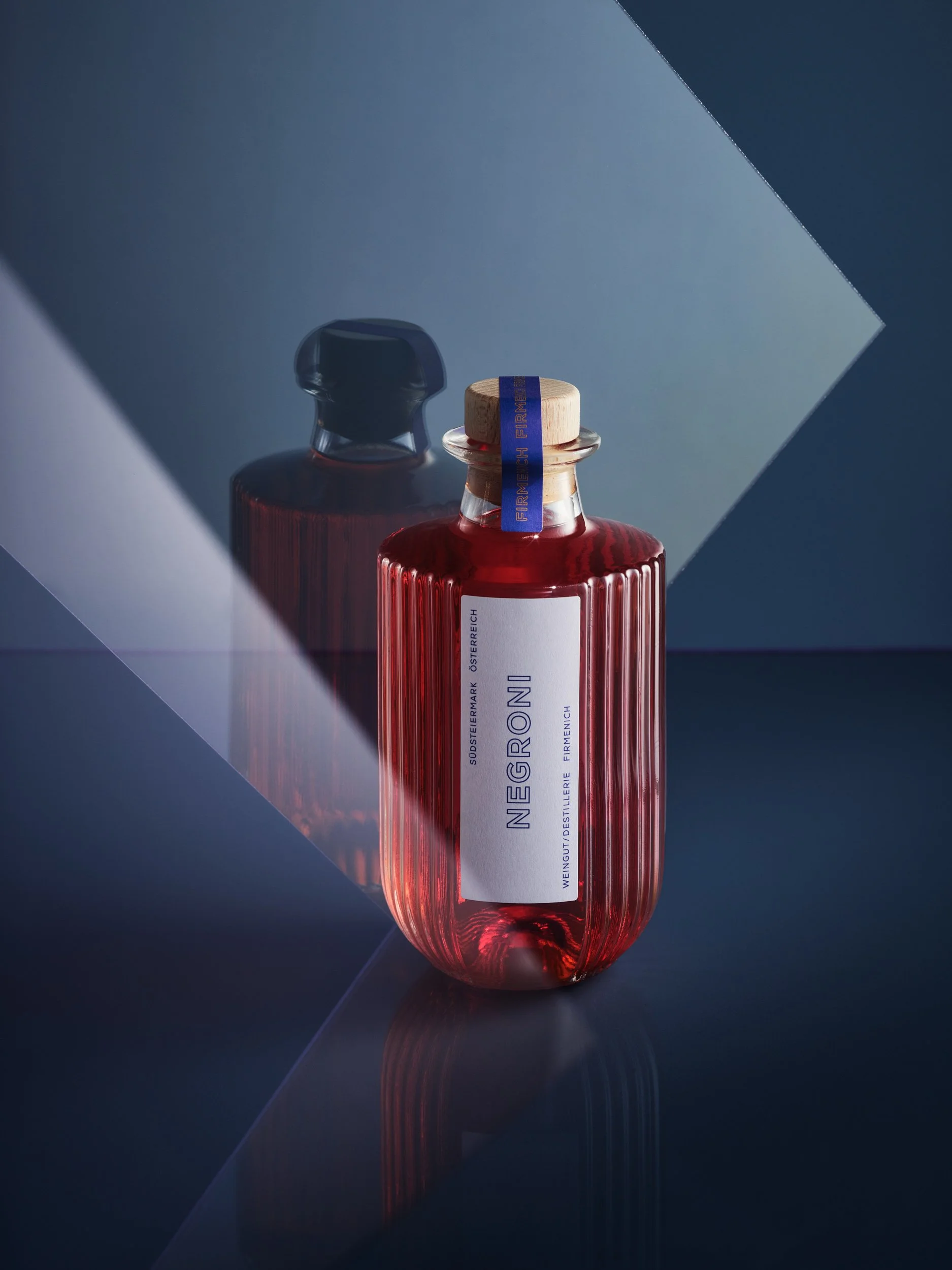 Eine rote Flasche mit Holzverschluss und weißem Etikett, die auf einer spiegelnden Oberfläche steht, im Hintergrund eine dunkle Flasche mit schwarzem Verschluss, beide im Schatten und Lichtreflexionen sichtbar.