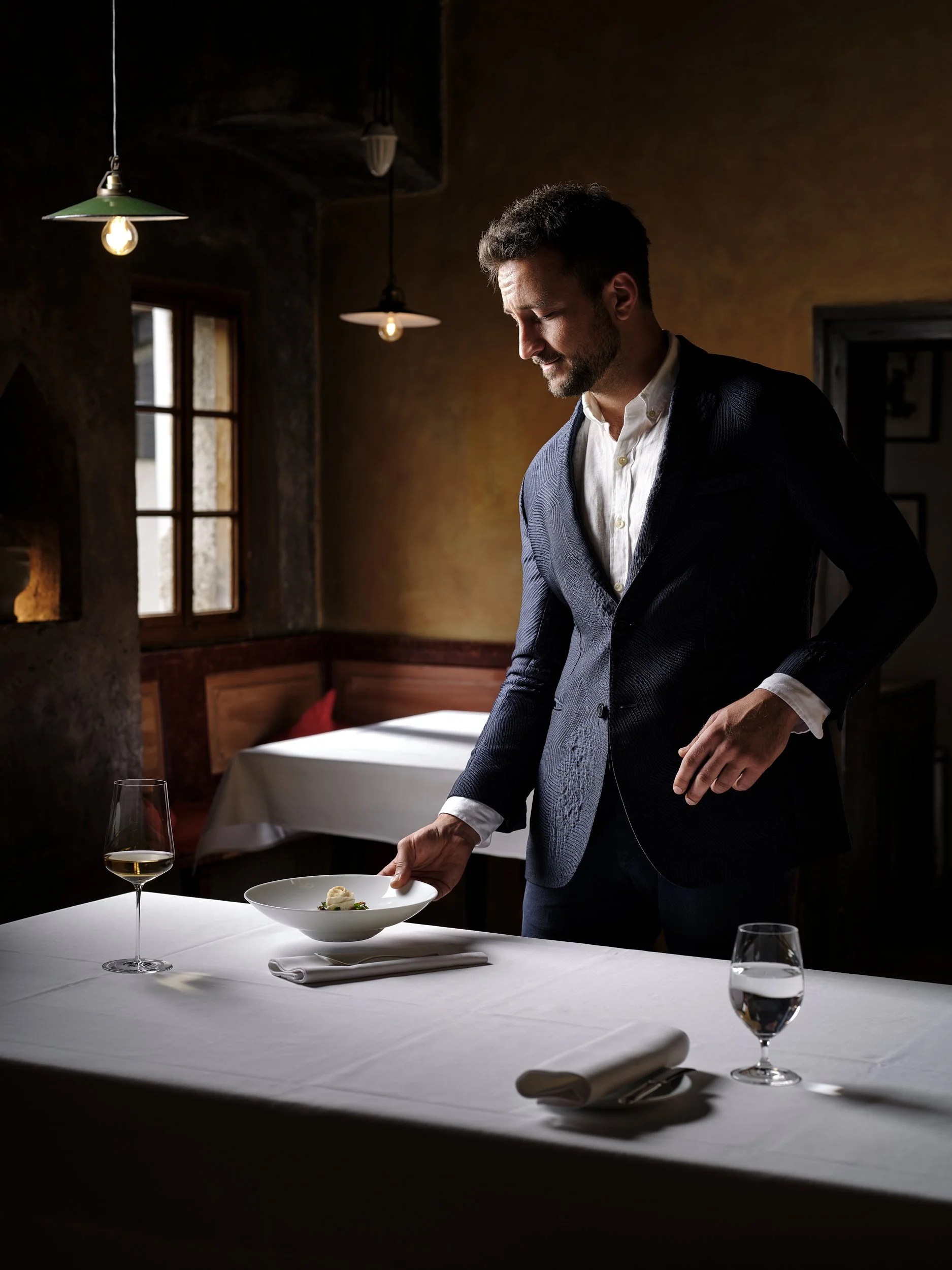 Ein Mann im Anzug steht in einem Restaurant mit gedämpfter Beleuchtung und hält einen Teller. Auf dem Tisch befindet sich ein Weinglas, ein Glas Wasser und Besteck auf einem gefalteten Tuch.