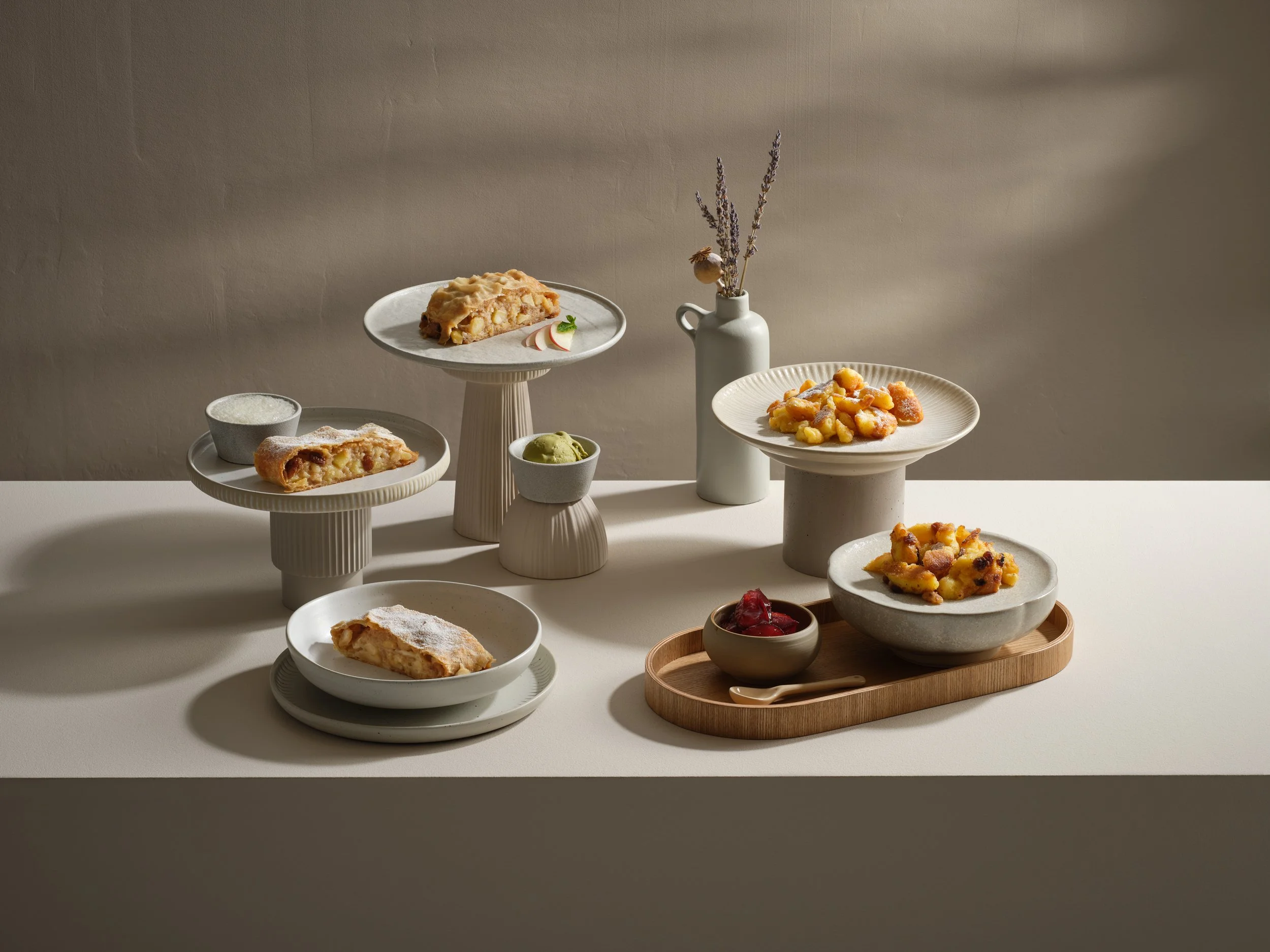 Verschiedene Kuchenstücke und Desserts auf weißen Tellern und Platten, einige mit Staubzucker, auf einer weißen Tischdecke, mit einer weißen Vase mit Lavendel und anderen Blumen im Hintergrund.