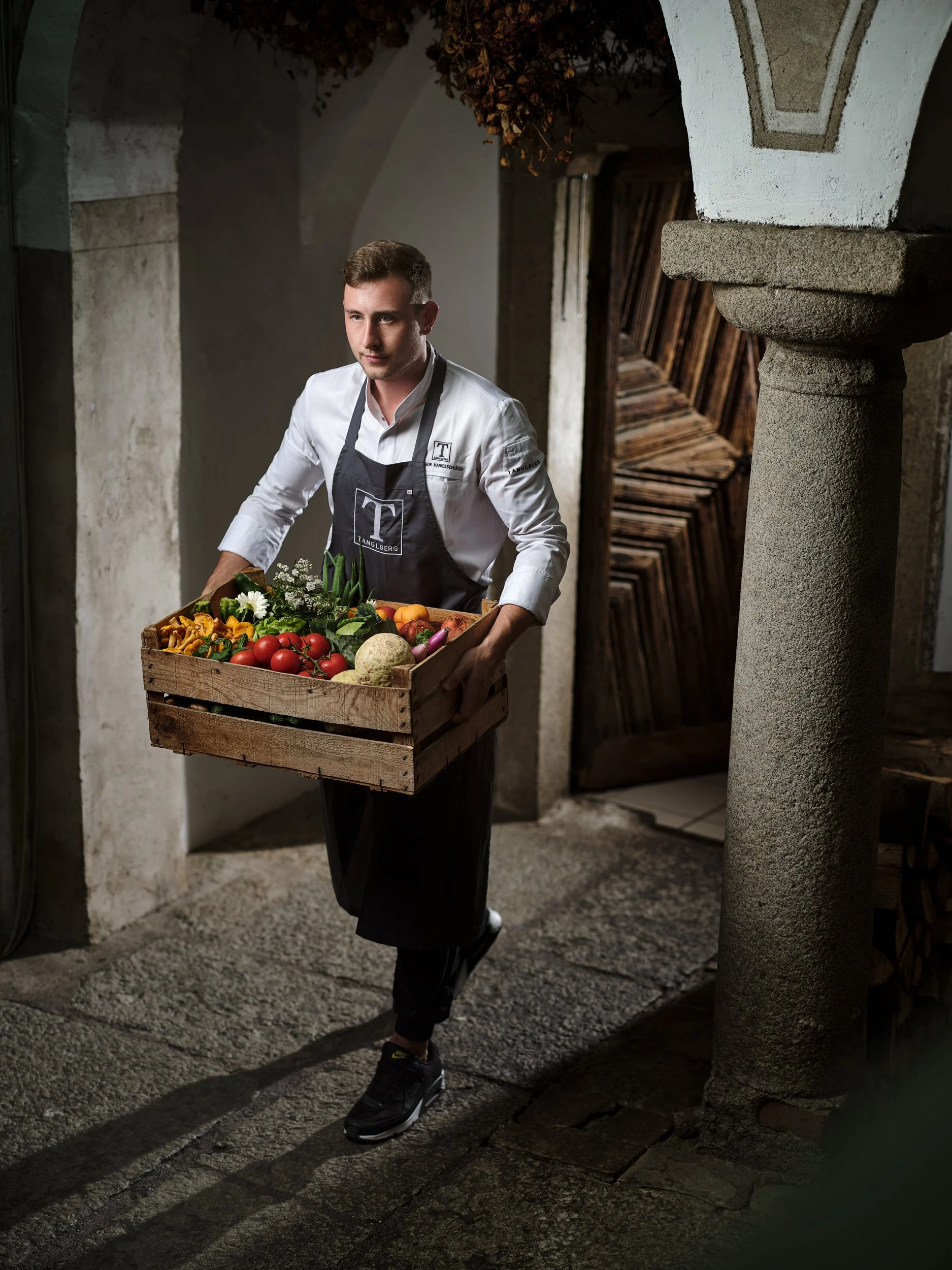 Ein Mann in einem weißen Hemd und schwarzer Schürze trägt eine Holzkiste voller frischem Gemüse und Obst. Er befindet sich in einem Durchgang mit steinernen Säulen und einem Holztor im Hintergrund.