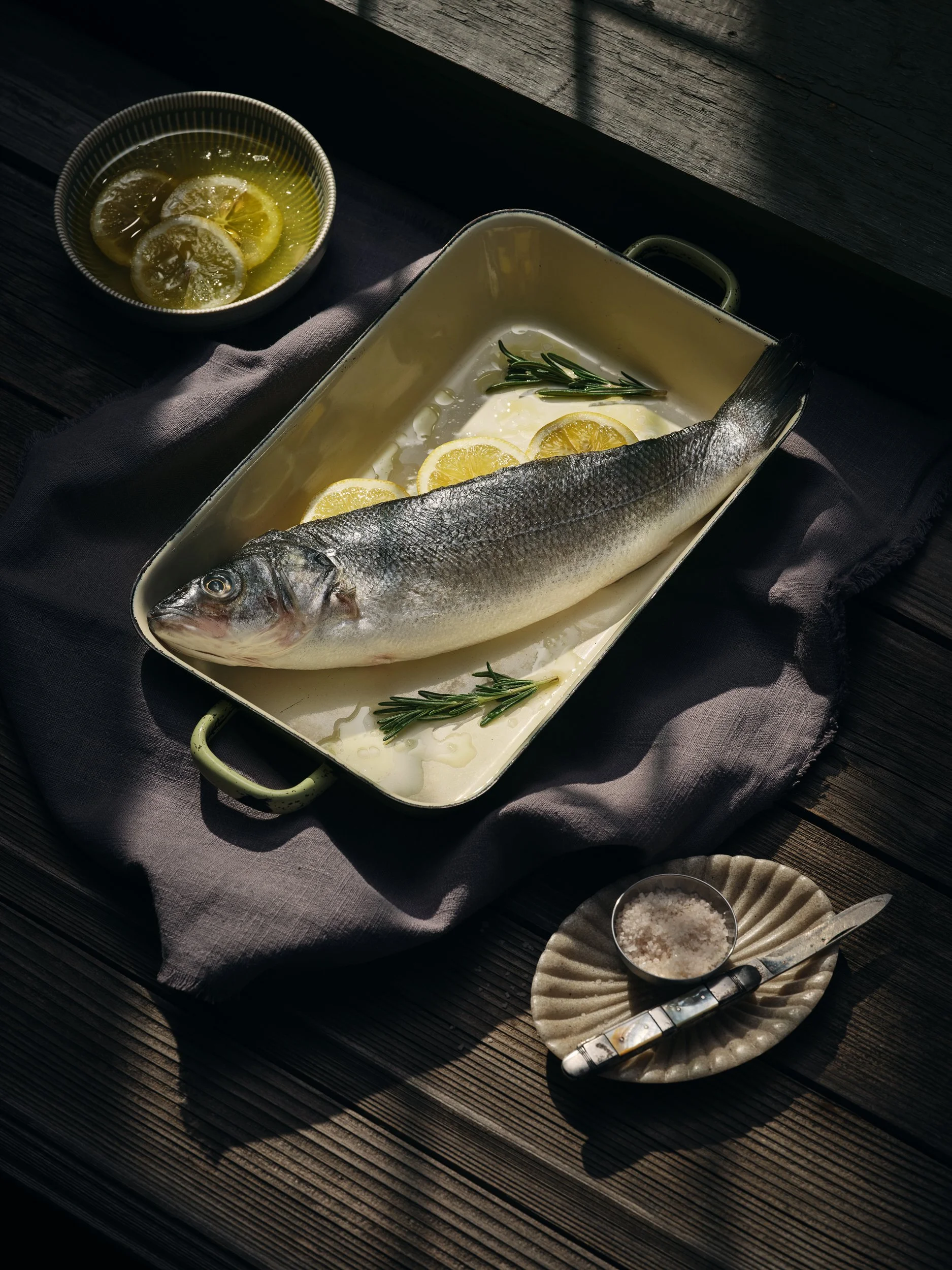 Fisch in einer Auflaufform mit Zitronenscheiben und Kräutern, daneben eine Schale mit Zitronenscheiben in Öl und ein Teller mit Salz und Messer auf Holztisch.