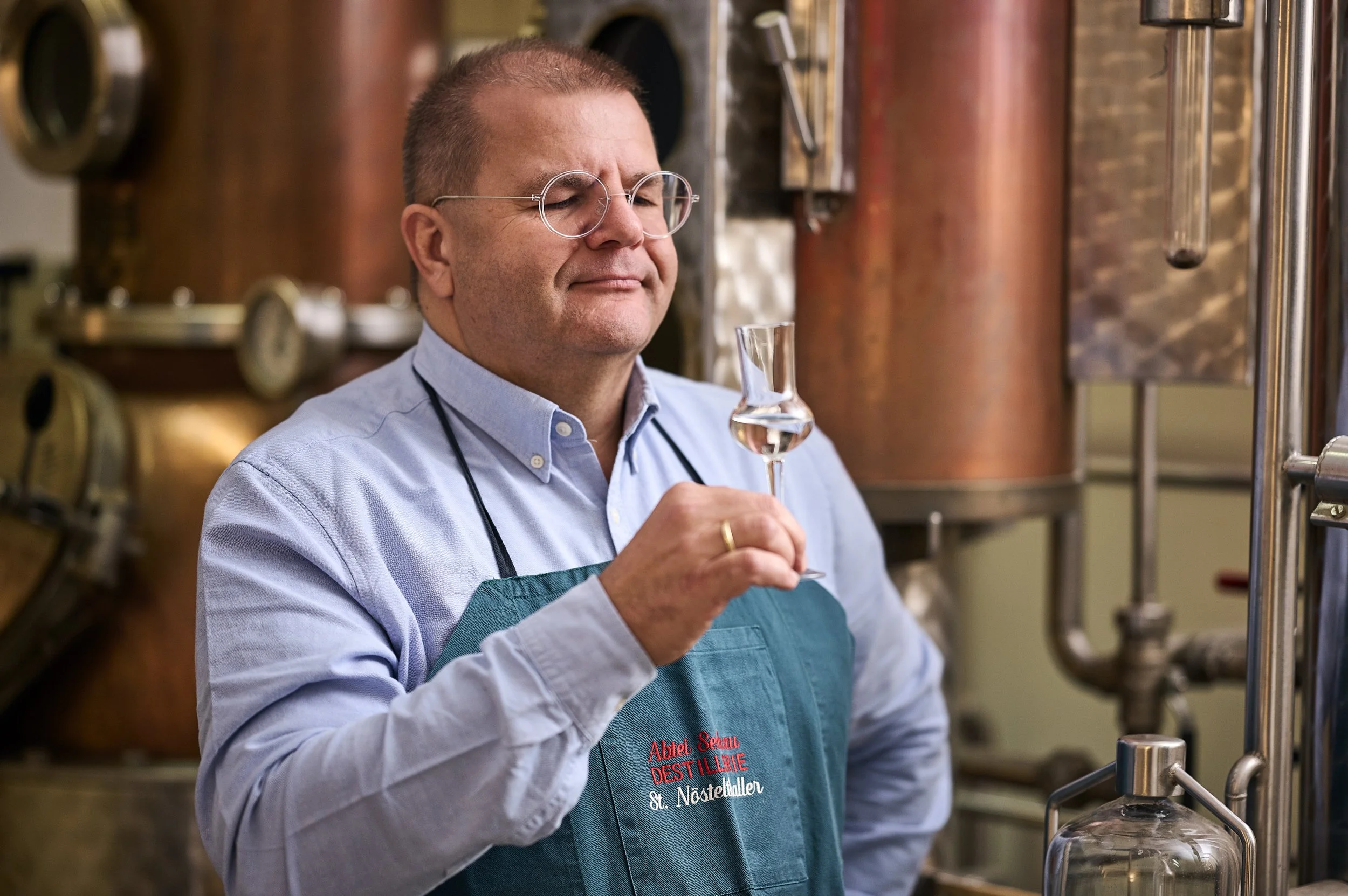 Ein Mann in Brille und blauer Kochschürze mit roten Aufdruck, der einen Wein im Keller hält und riecht.