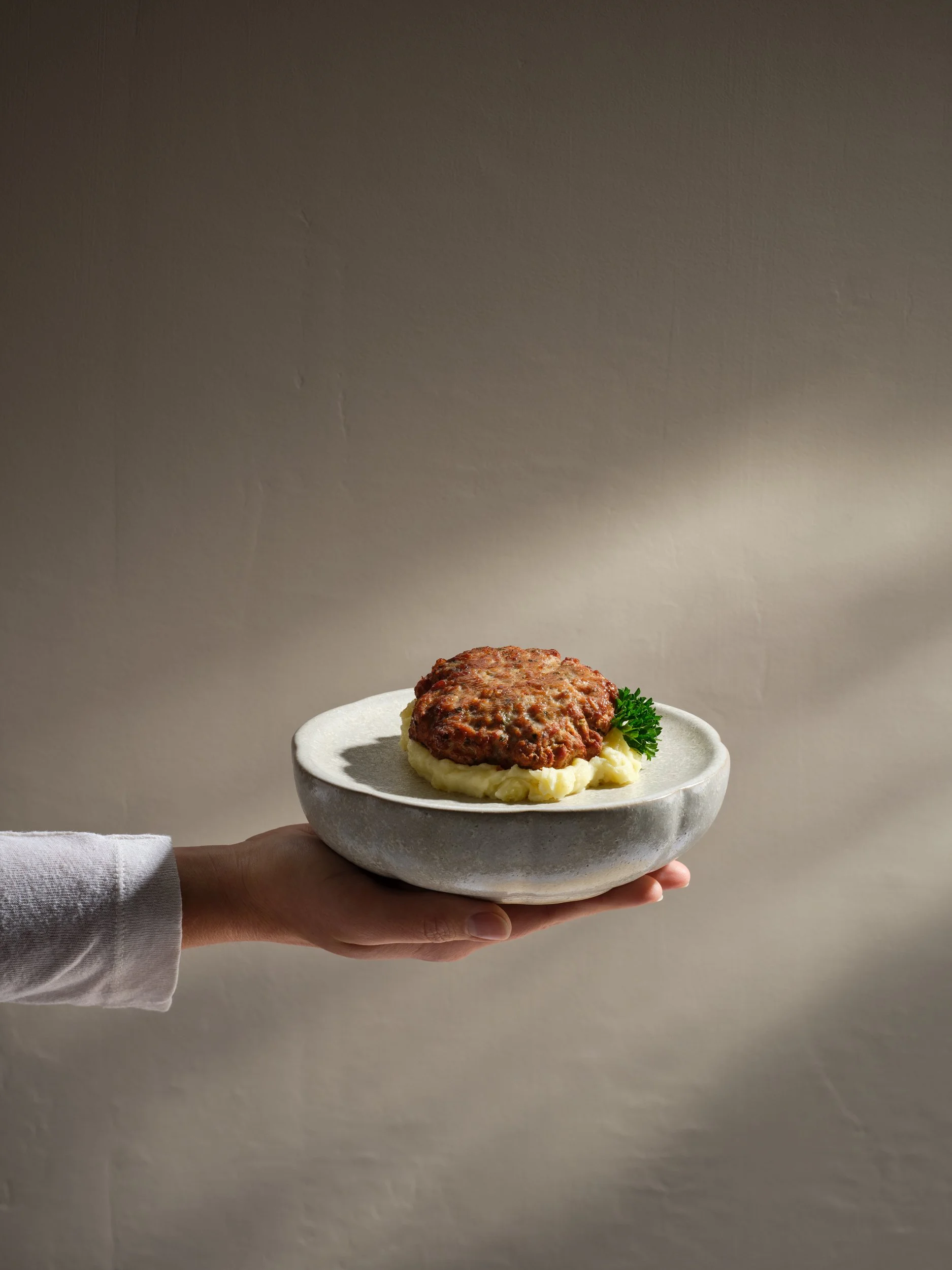Eine Hand hält einen Teller mit einem Hamburger-Pattie, Kartoffelpüree und Petersilie.