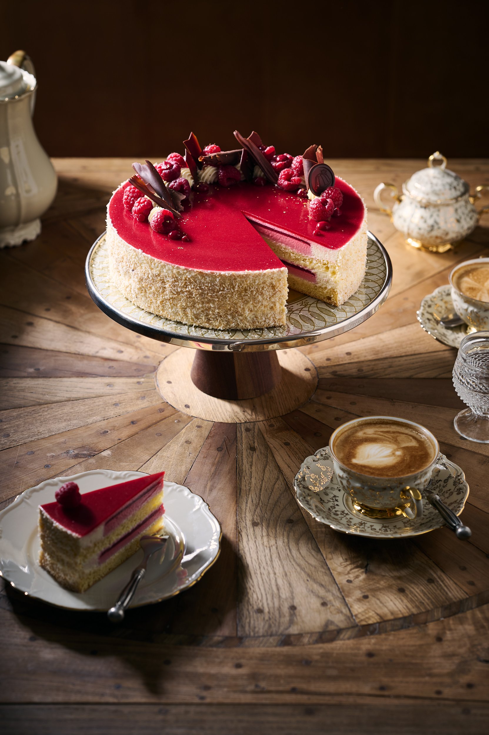 Ein roter Erdbeerkuchen mit Beeren und Schokoladenspänen, daneben eine Tasse Cappuccino, auf einem Holztisch.