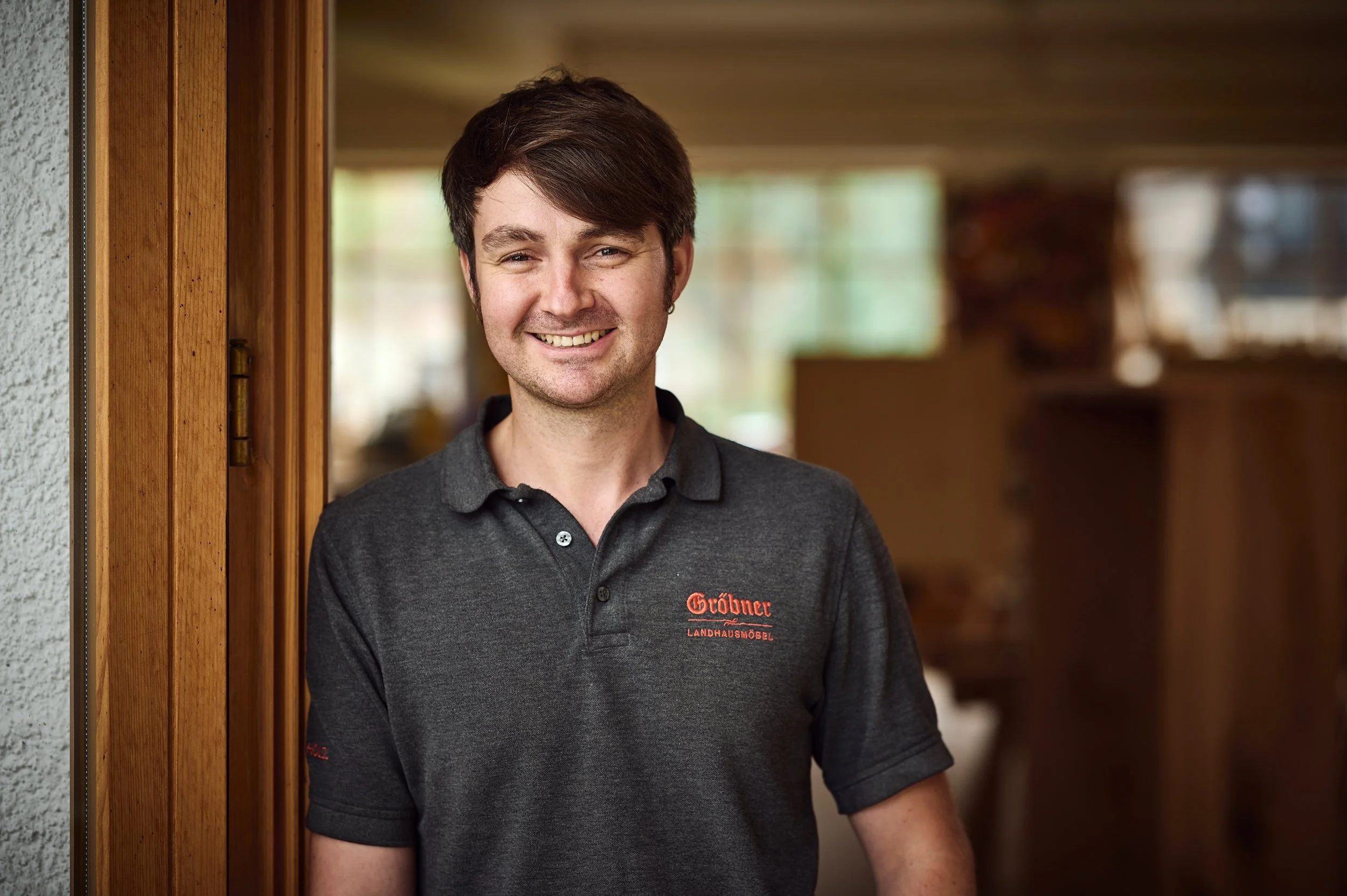 Ein junger Mann mit dunklen Haaren und einem grauen Poloshirt, das das Logo einer Landhausmöbelfirma trägt, steht vor einer Holzwand in einem Raum mit viel natürlichem Licht, im Hintergrund unscharf.