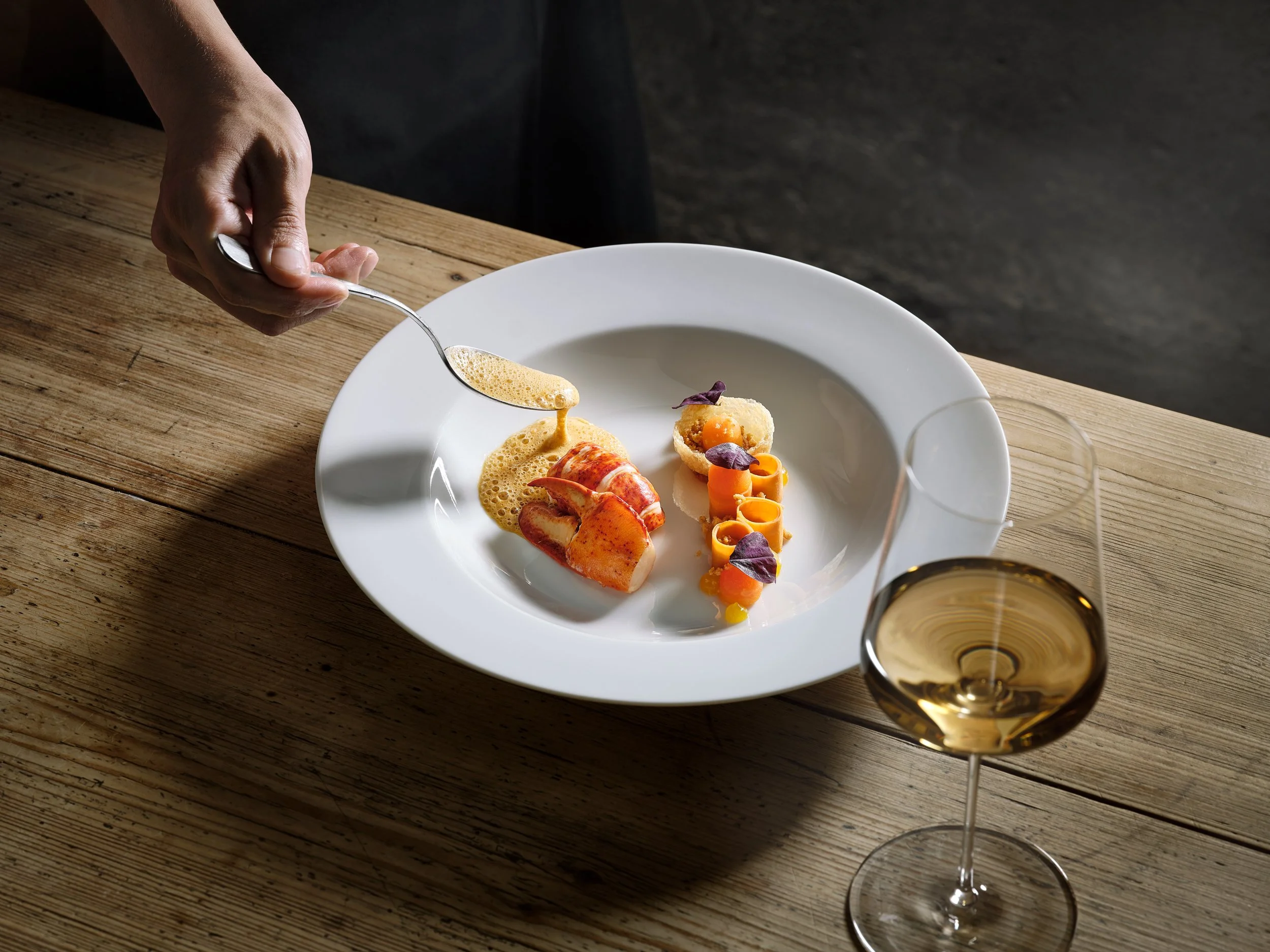 Gourmetgericht auf hellem Holz-Tisch. Eine Hand gießt Soße über ein kunstvoll angerichtetes Gericht mit Meeresfrüchten, Karotten-Röllchen und Kräutern in einem weißen Teller. Ein Glas Weißwein steht daneben.