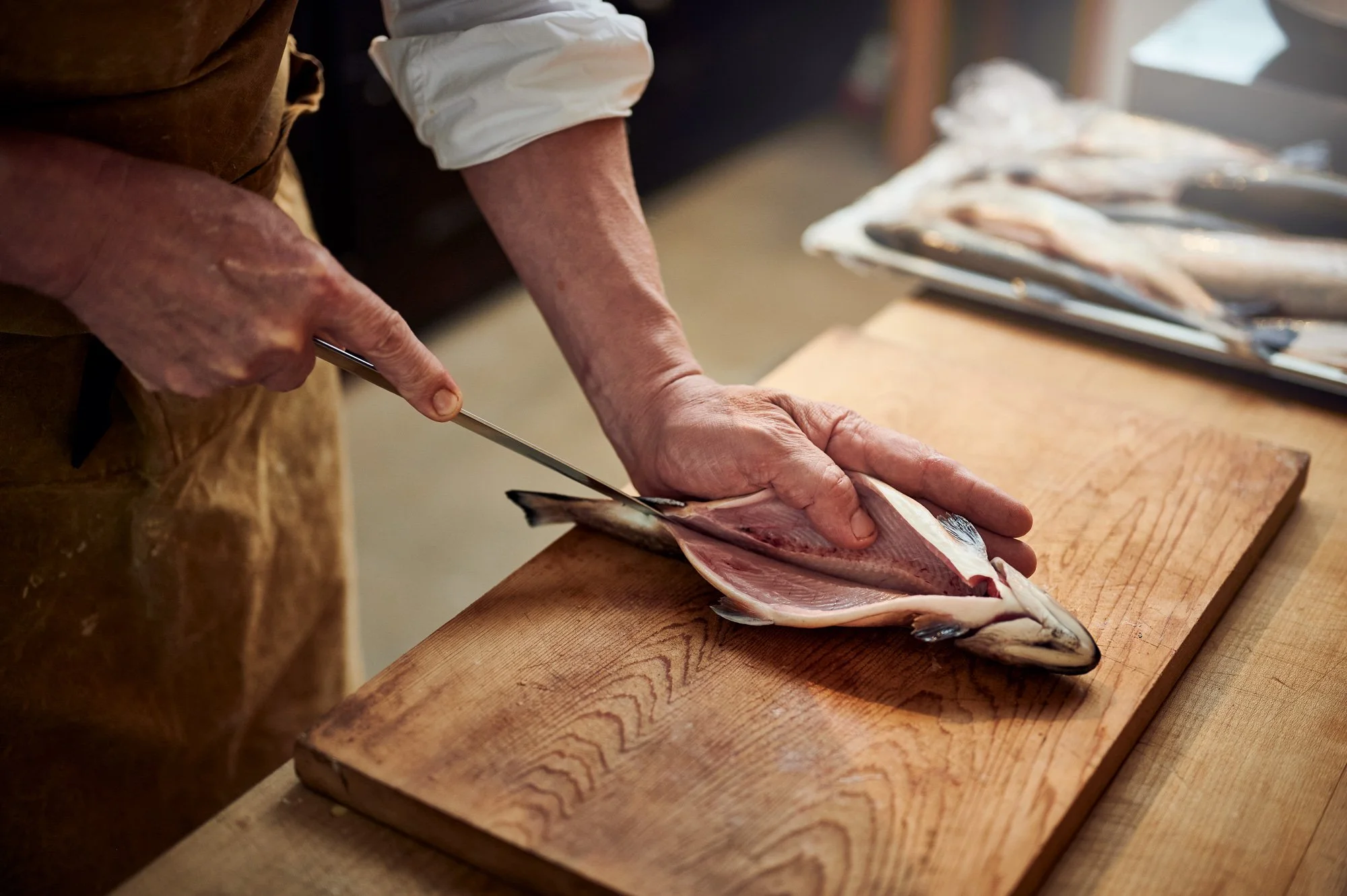 Nahaufnahme eines Handwerks, das eine Fischfiletierung auf einem Holzbrett durchführt. Es sind mehrere gefischte und filetierte Fische auf einem Tablett im Hintergrund.