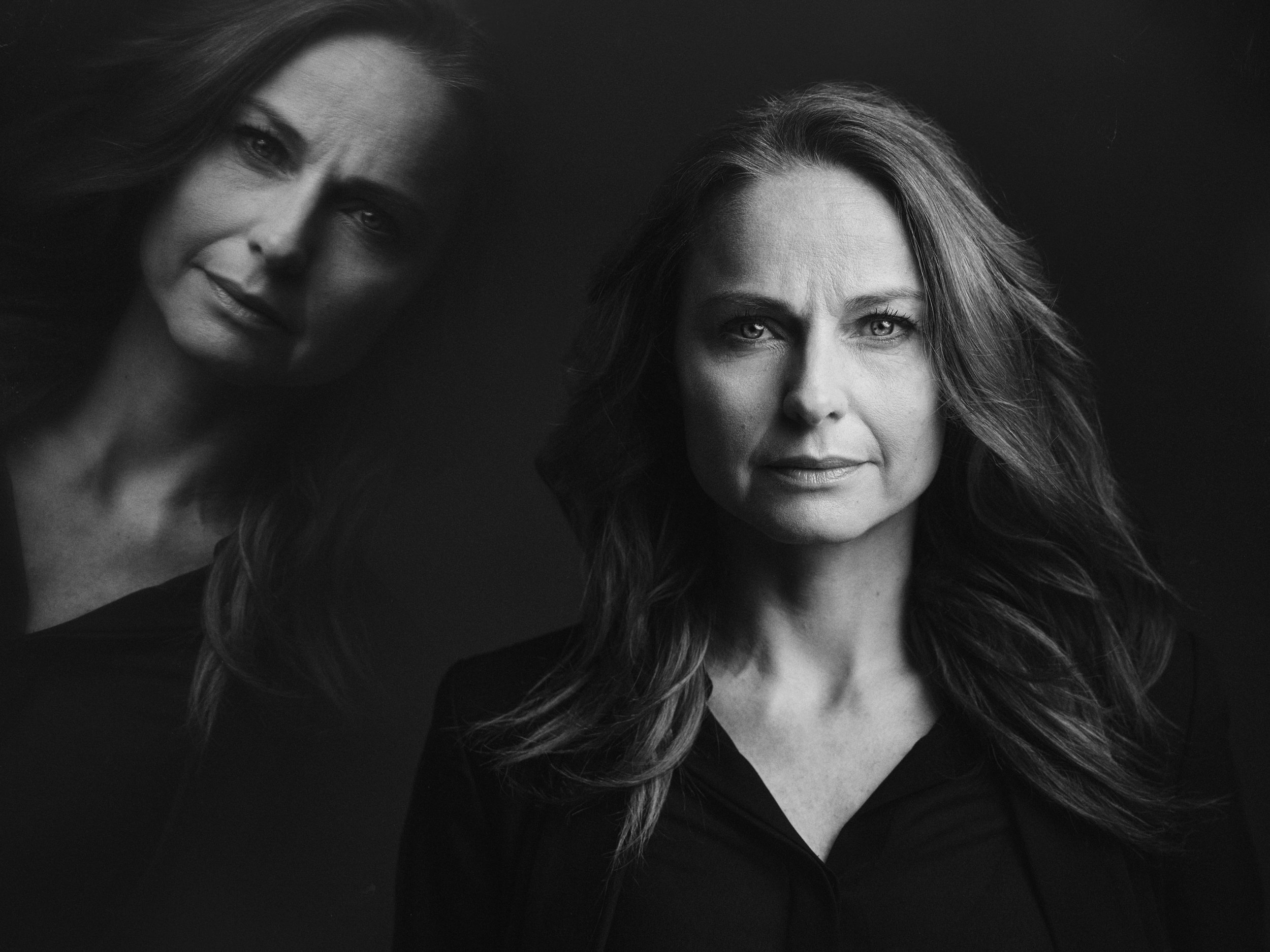 Schwarz-weiß Porträt einer Frau mit langem Haar und ernstem Gesichtsausdruck, mit einer spiegelnden Reflektion links im Bild.