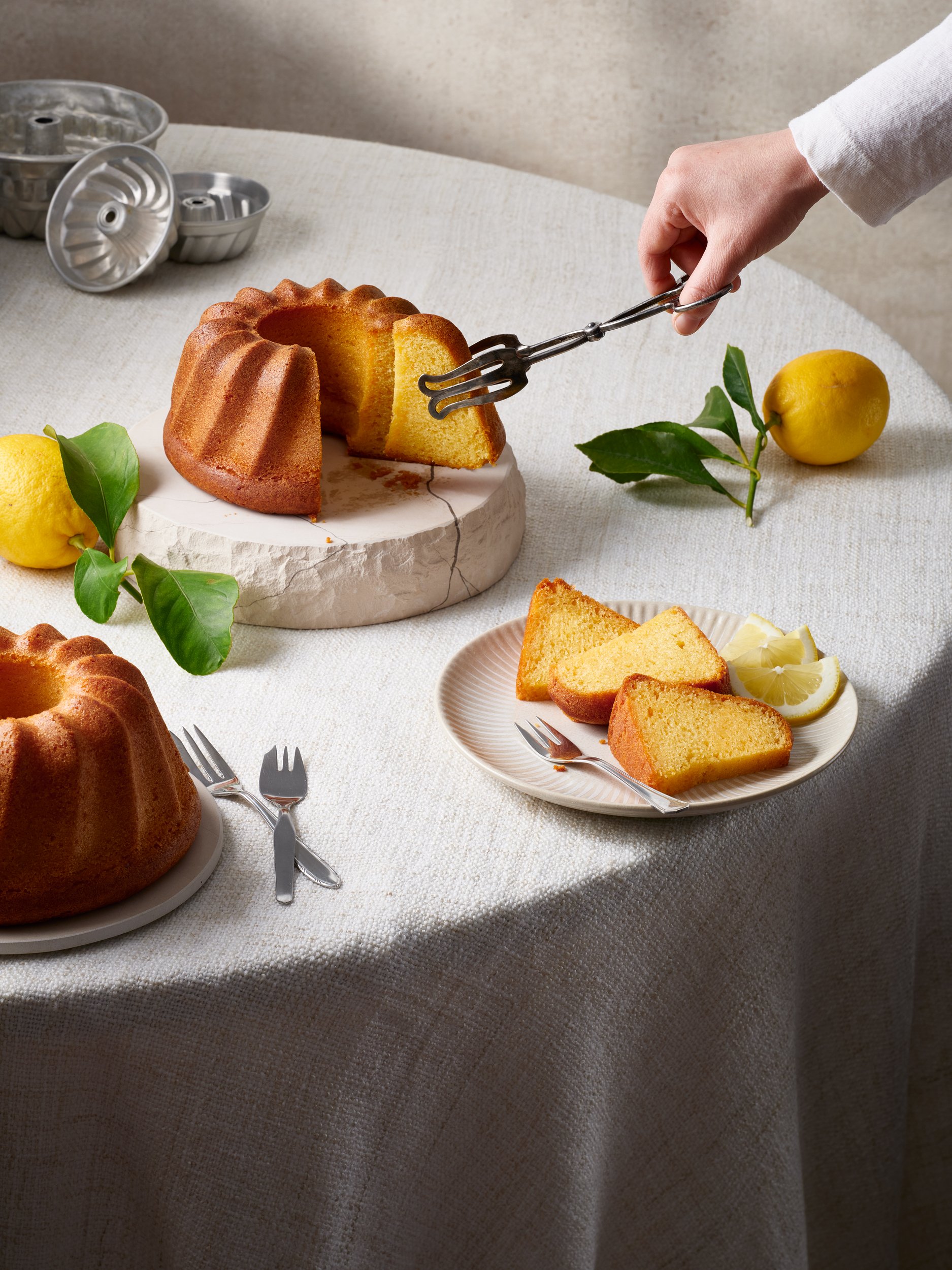 Ein gedeckter Tisch mit goldgelbem Gugelhupf, Zitronenscheiben, Käse und Zitronen, sowie einer Hand, die ein Stück Kuchen mit einer Gabel schneidet.