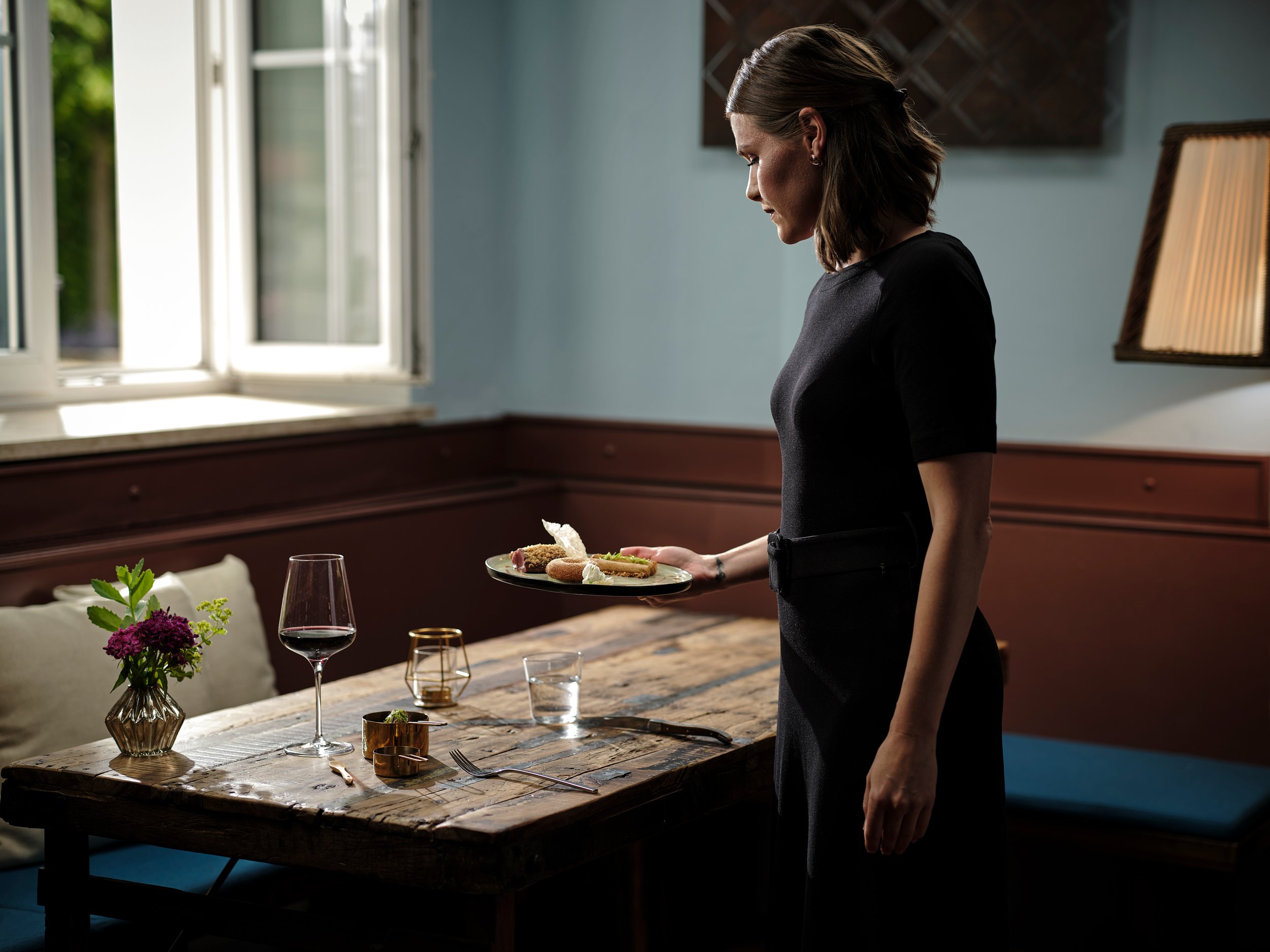 Eine Kellnerin hält einen Teller mit Essen in einem Restaurant, während sie neben einem rustikalen Holztisch steht, auf dem Gläser, Besteck und eine Vase mit Blumen stehen.