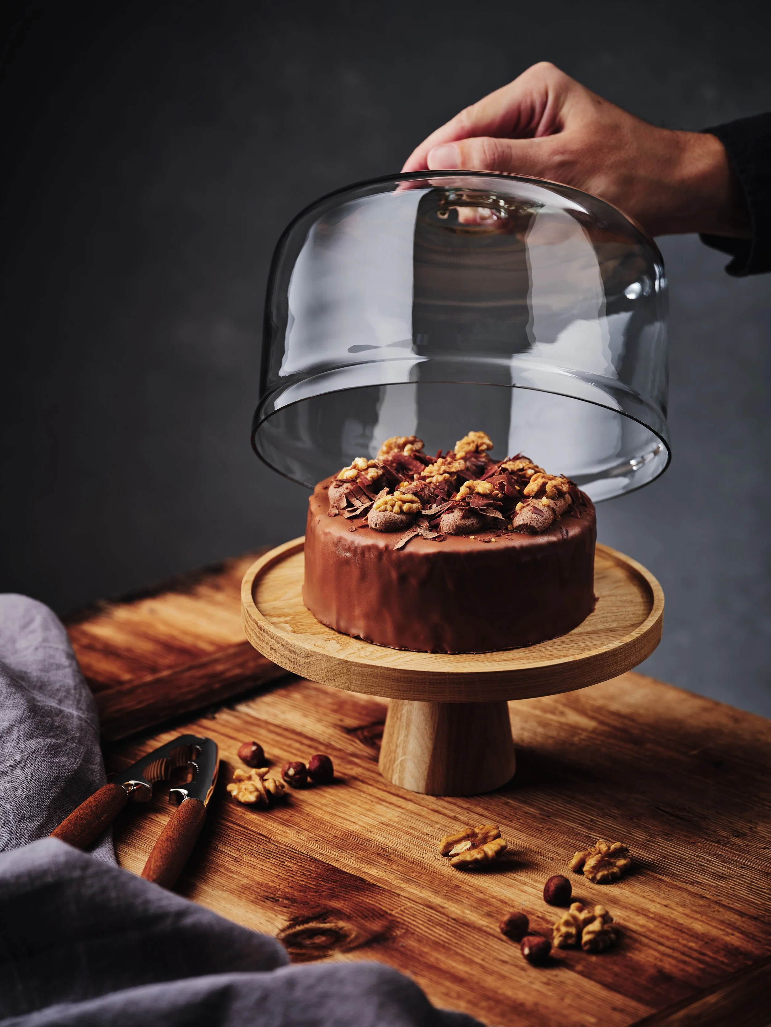 Hand hebt Glashaube von einem Walnuss-Schokoladenkuchen auf Holzständer, mit Nüssen und Nussknacker auf Holztisch.