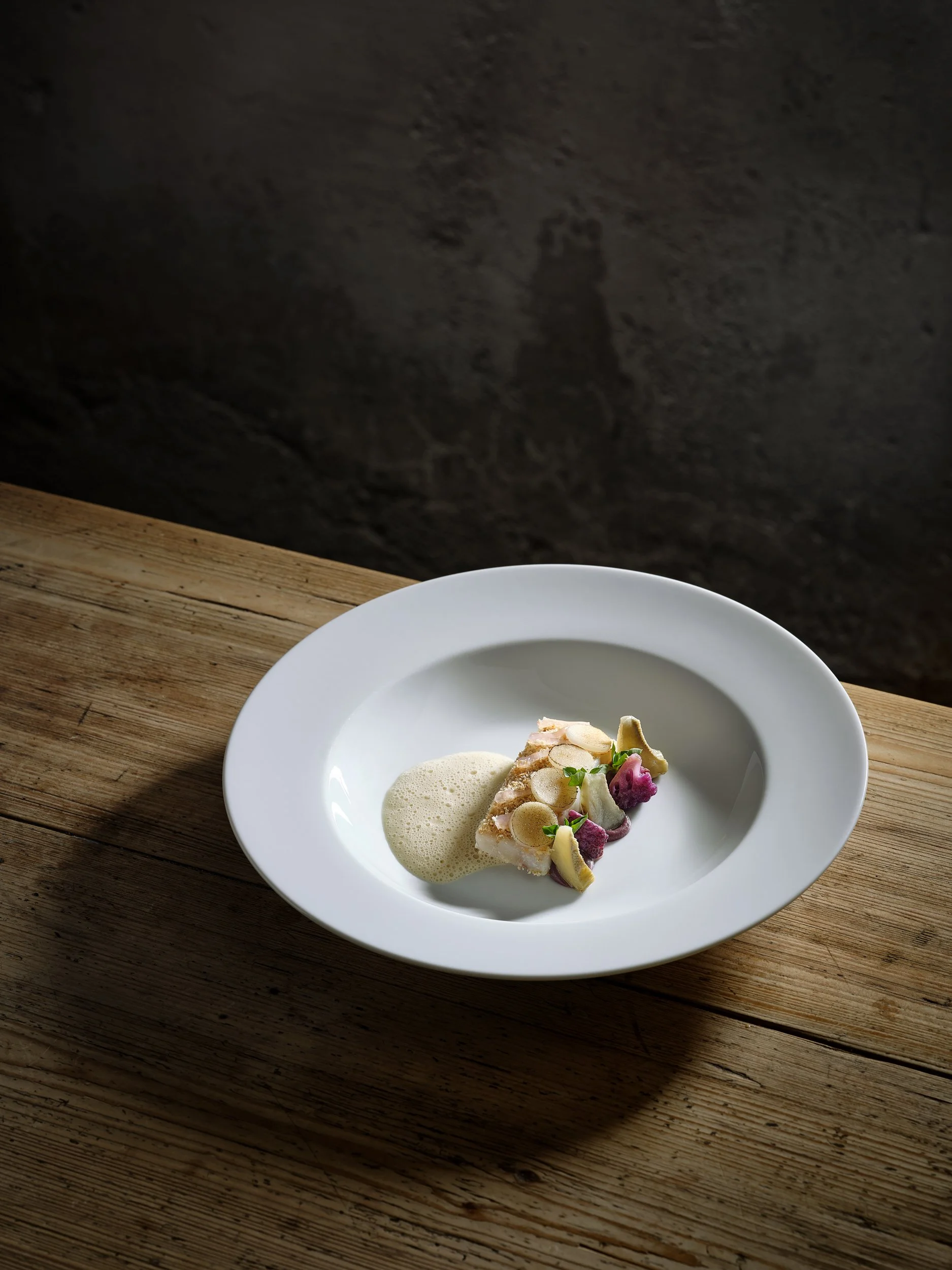 Gourmetgericht auf einem weißen Teller mit Schaum, Gemüse und Fischfilet. Auf einem Holzttisch arrangiert.