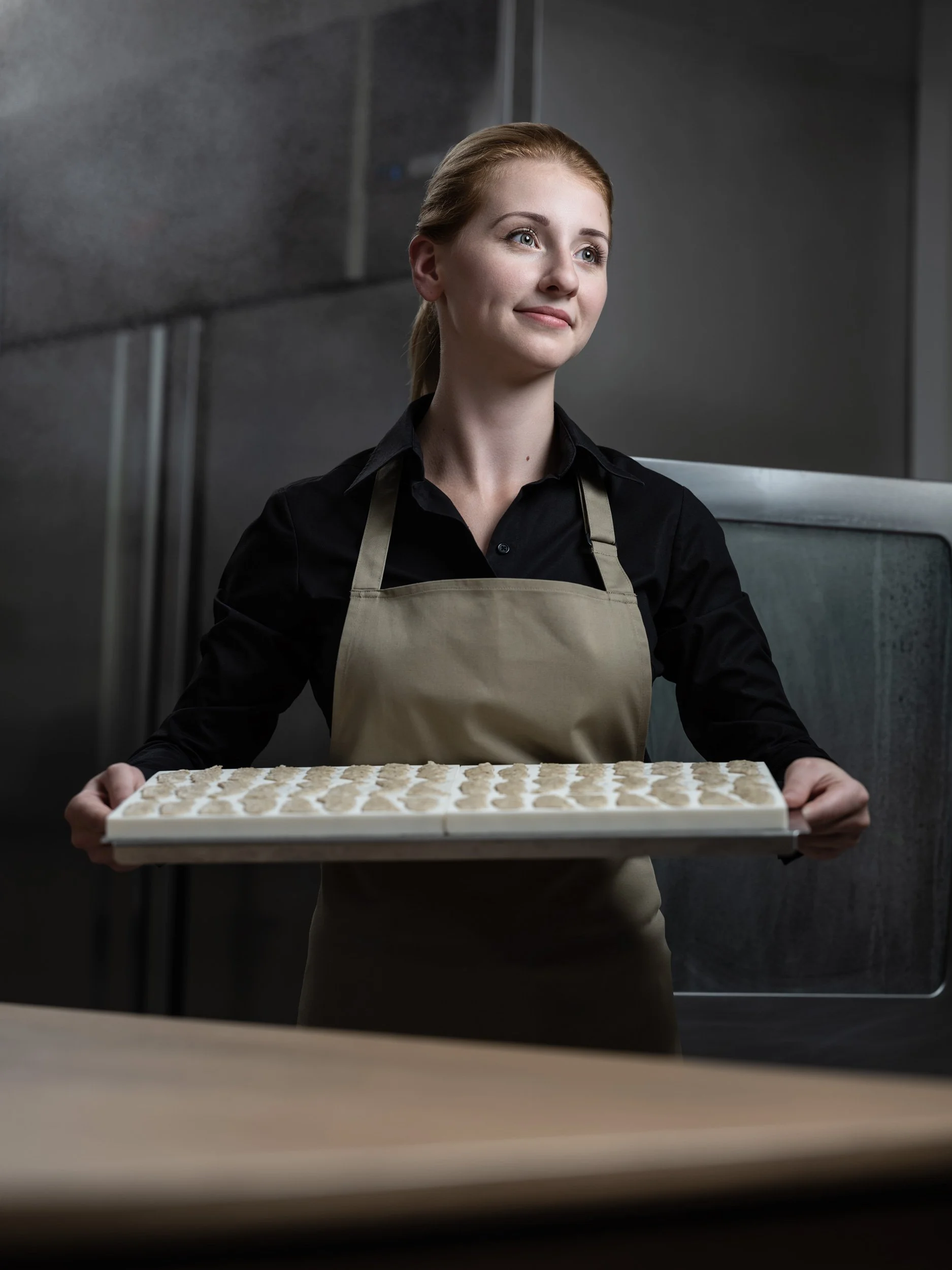 Frau in schwarzem Hemd und Schürze hält ein Tablett mit kleinen Formen in einem industriellen Küchenumfeld.