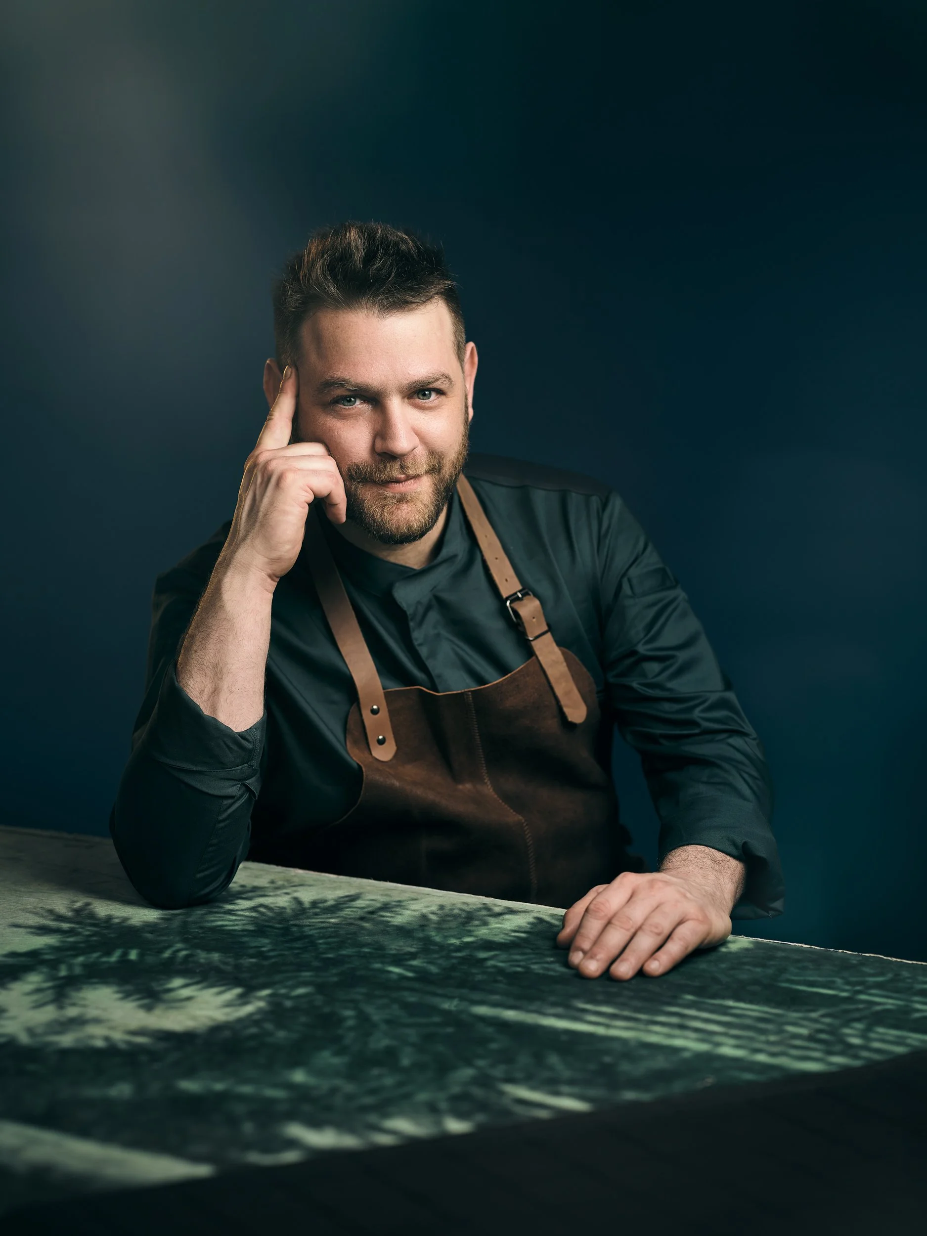 Ein Mann mit dunklem Hemd und brauner Leder-Schürze sitzt an einem Tisch und betrachtet eine Karte oder ein Bild, vor einer dunklen, einfarbigen Wand.