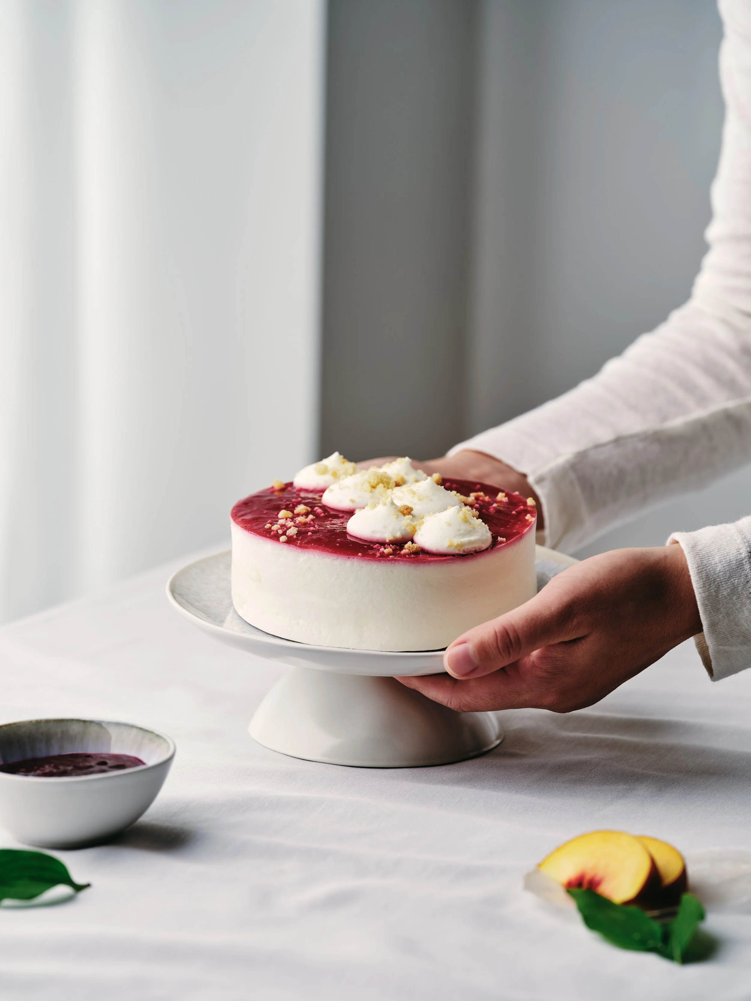 Person hält einen runden Kuchen mit rotem Belag und Sahnehäubchen auf einem weißen Kuchenteller. Daneben steht eine Schale mit Sauce und ein paar Pfirsichscheiben auf einem Tisch mit weißem Tischtuch.