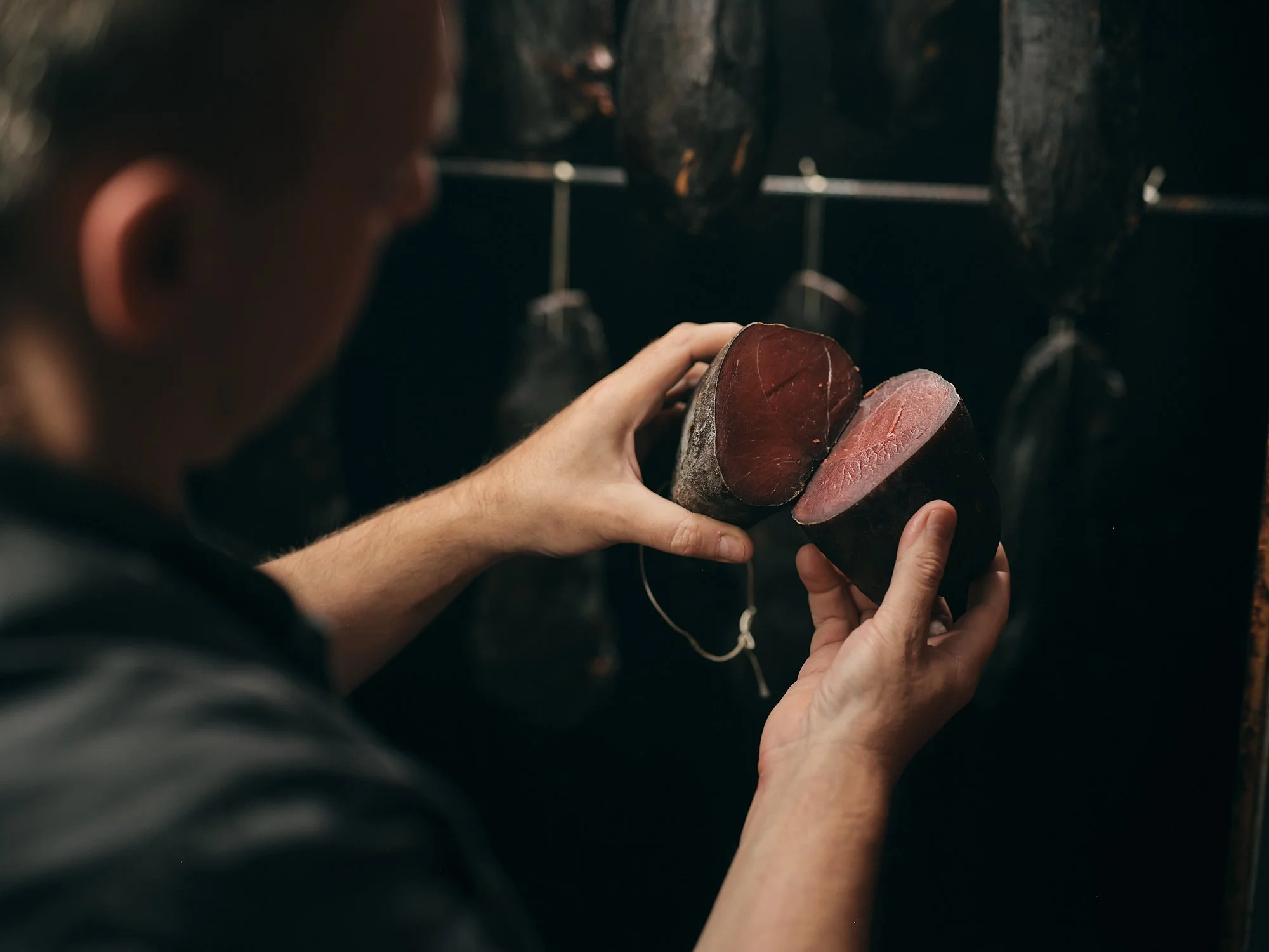 Eine Person hält ein Stück rohen Fisch, vermutlich Thunfisch, vor schwarzen, getrockneten Fischen, die an einer Wand hängen.