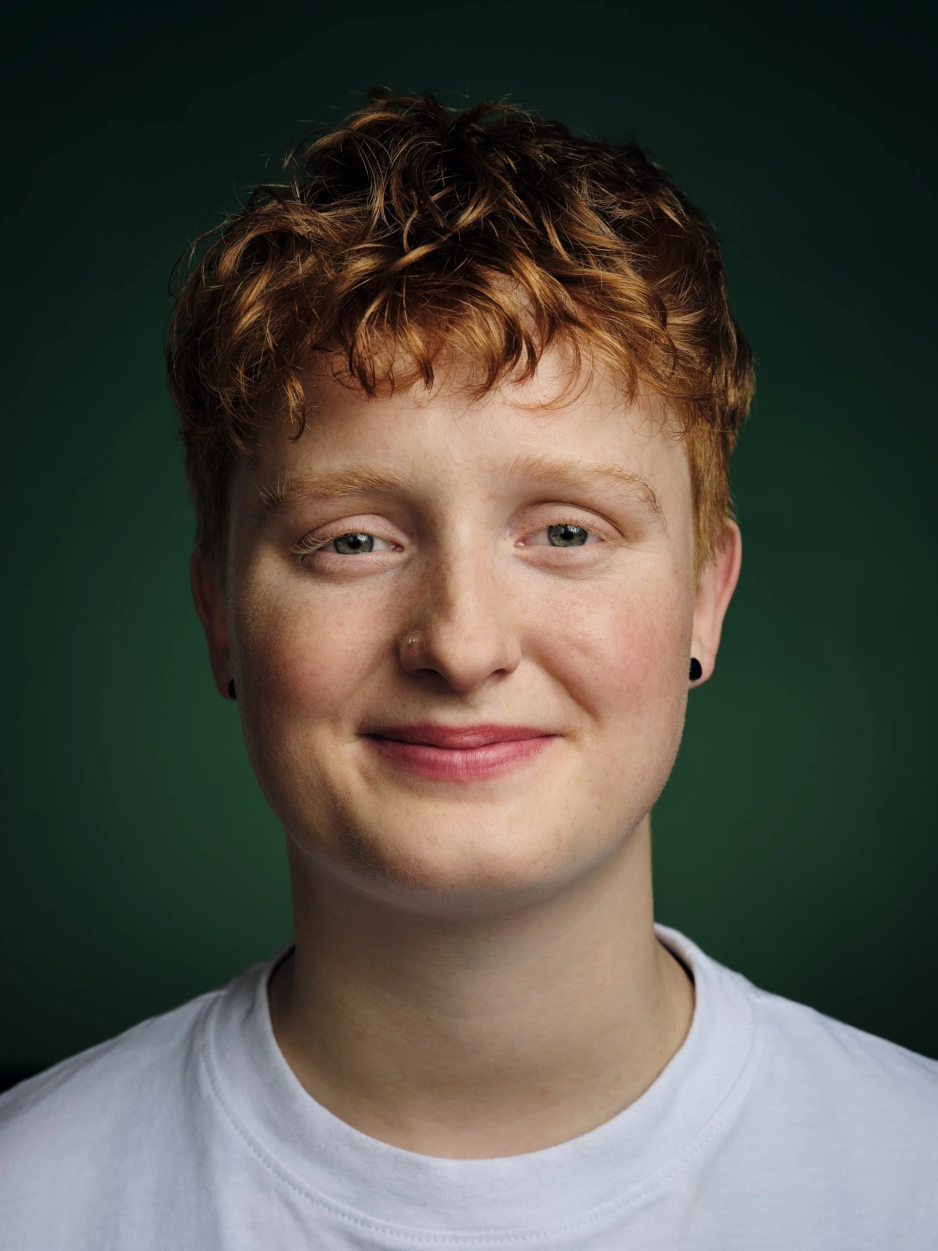 Nahaufnahme eines lachenden jungen Mannes mit roten, lockigen Haaren und blauen Augen, trägt ein weißes T-Shirt vor einem dunklen, grünen Hintergrund.