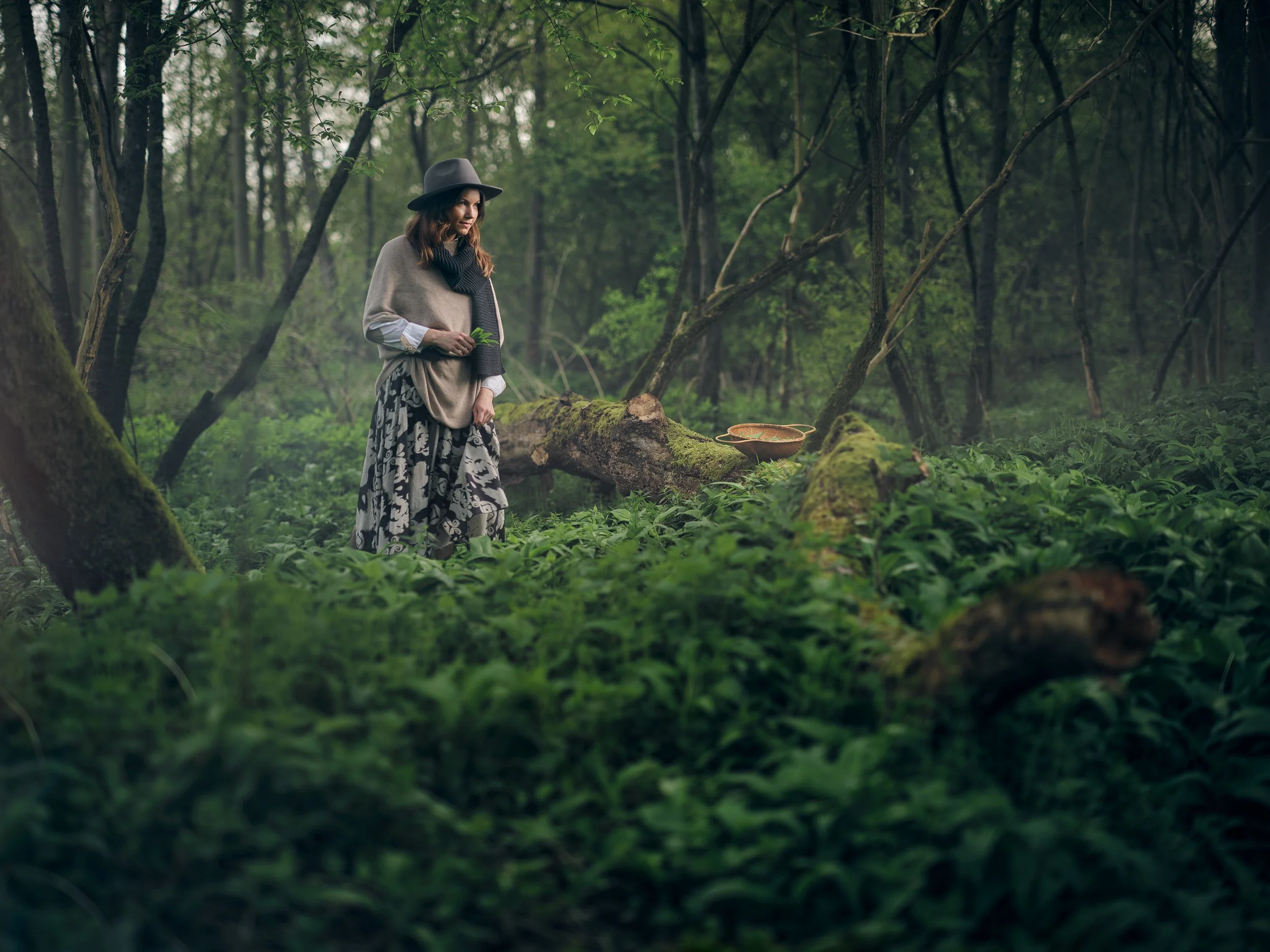 Eine Frau steht in einem grünen Wald voll mit Bärlauch, trägt einen Hut und Schal, und hält Bärlauch Pflanzen in der Hand. Im Hintergrund ist ein umgefallener Baumstamm mit Pilzen zu sehen. Der Wald ist dicht und neblig.