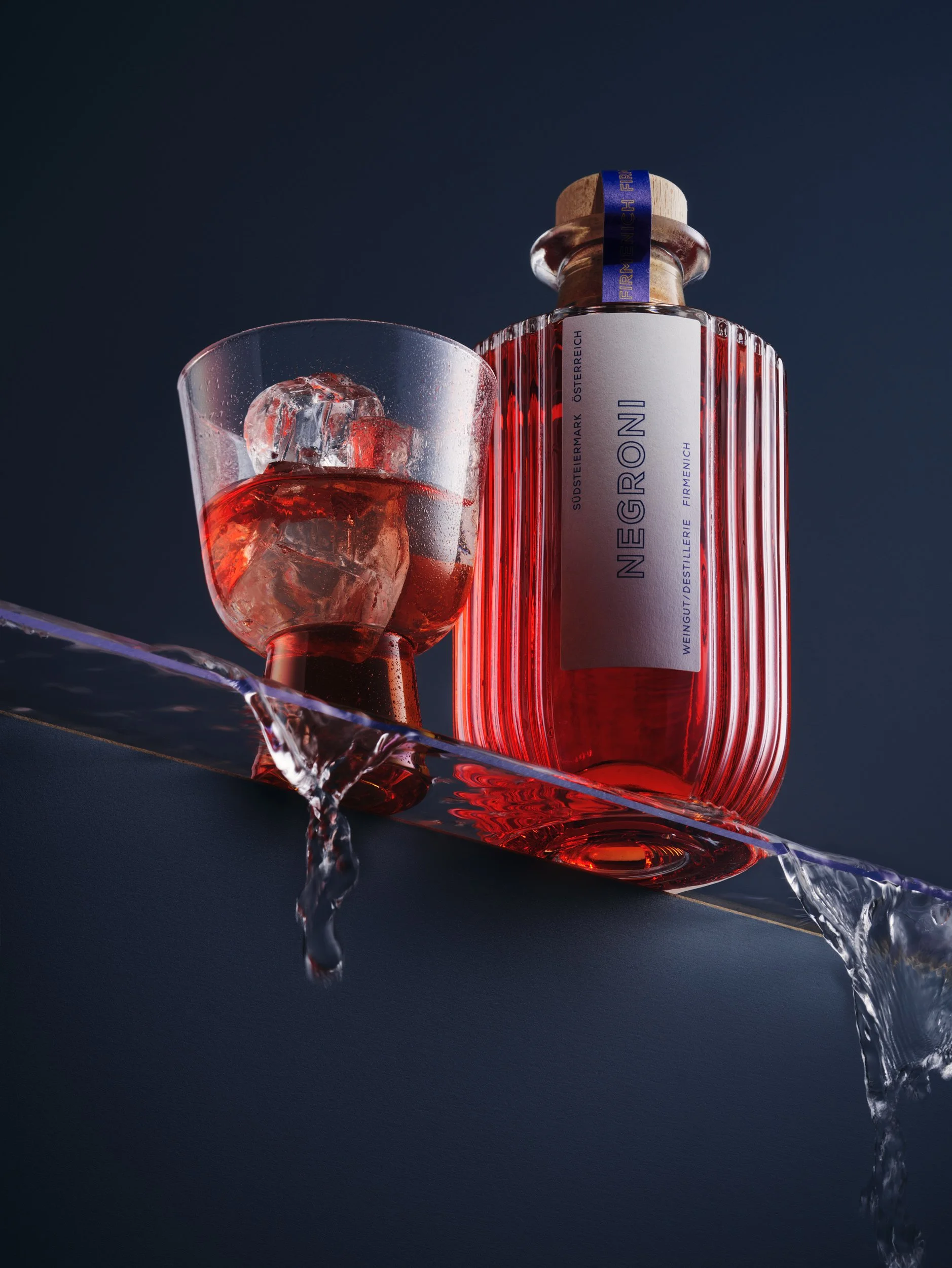 Eine Flasche Negroni neben einem Glas Negroni mit Eiswürfeln. Das Glas steht auf einer dunklen Oberfläche, der Hintergrund ist dunkelblau.