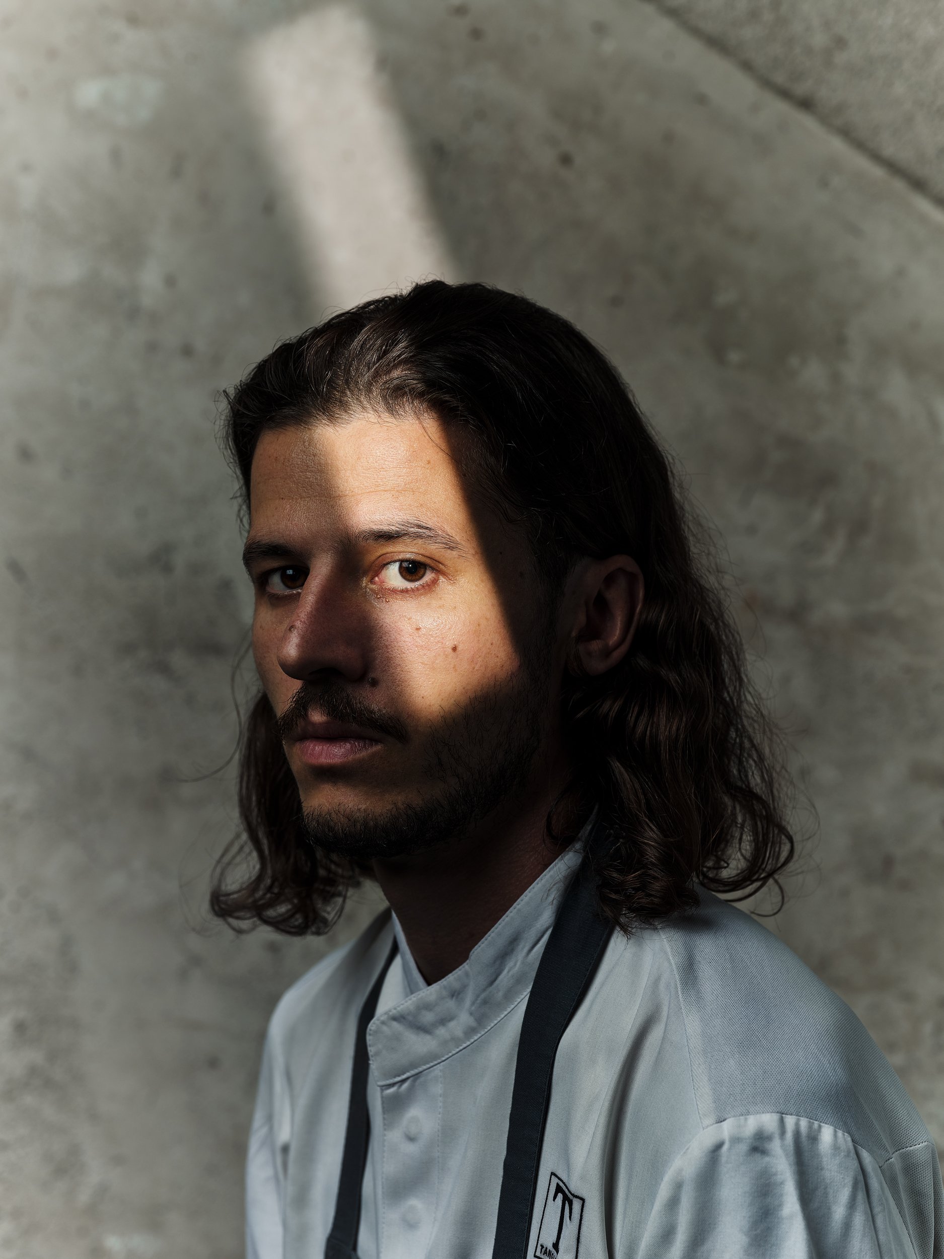 Ein Mann mit langen Haaren in einem Kochanzug, teilweise im Schatten, vor einem grauen Hintergrund.